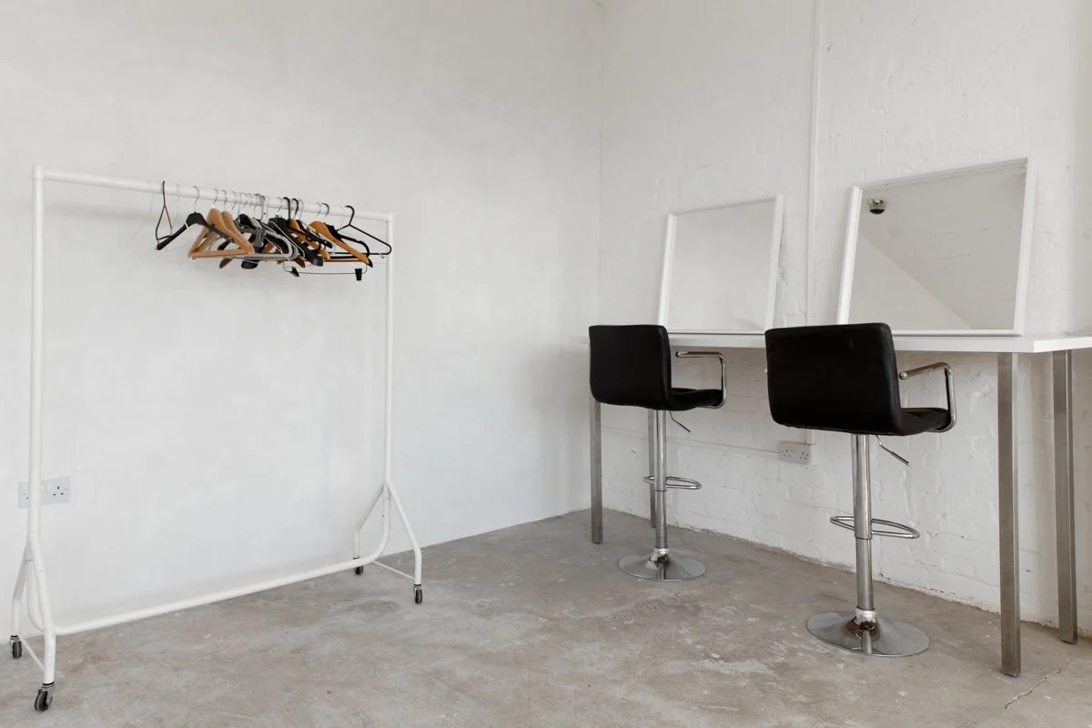 White room with two black chairs in front of mirrors, white table, and clothing rack with a few hangers and clothes.