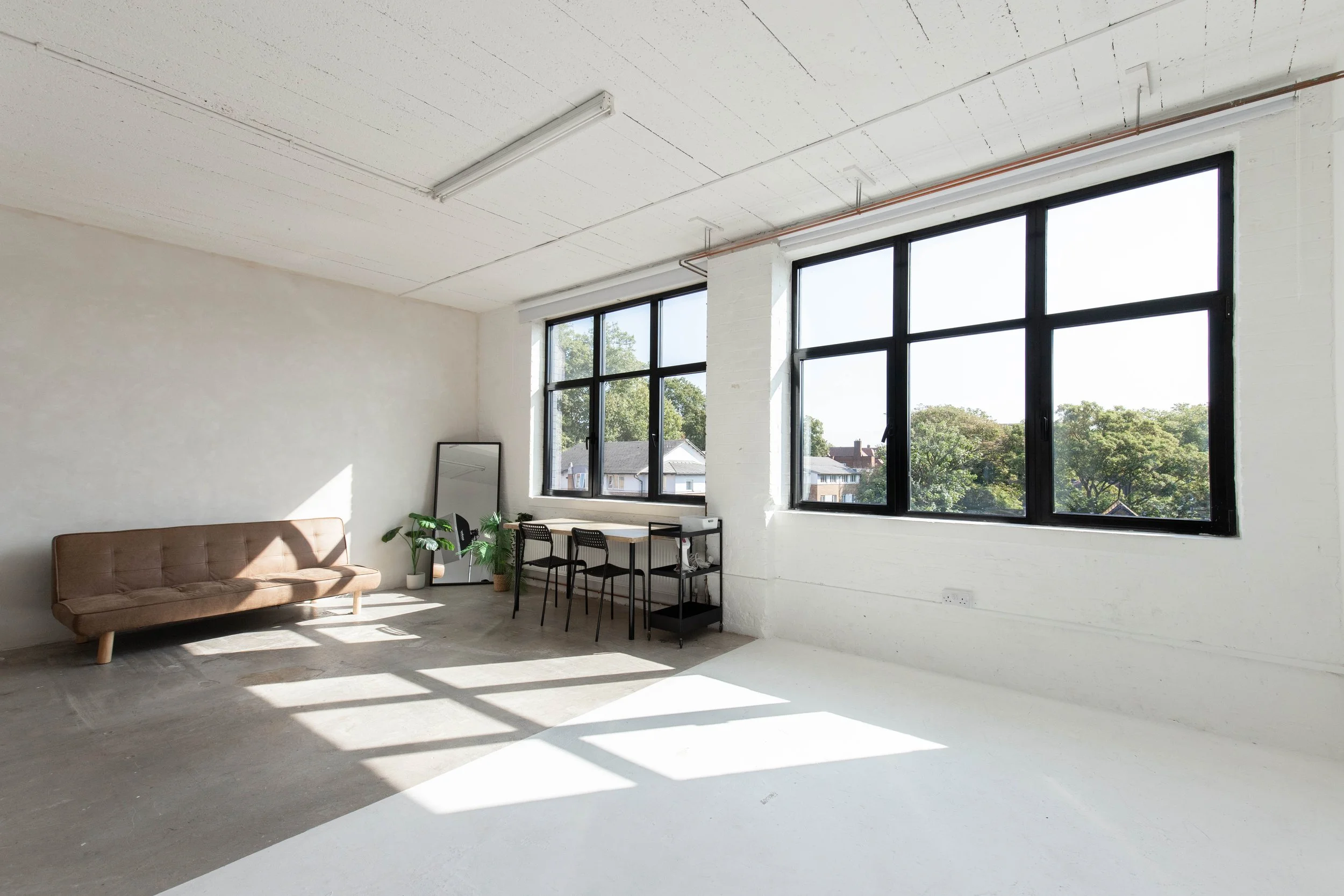 Bright, minimalist living space with large black-framed windows, a tan sofa, a black-framed mirror, a small table with black chairs, and potted plants, illuminated by natural sunlight.