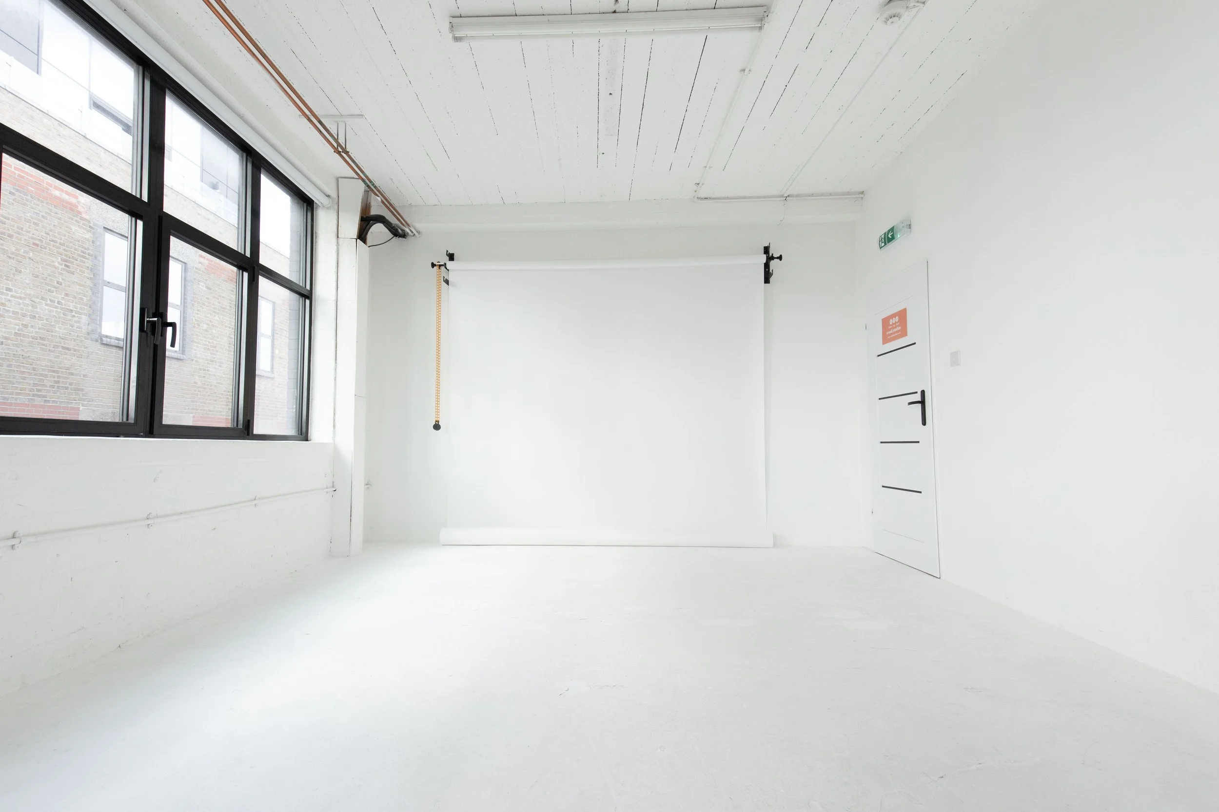 Empty photography studio with large windows, white walls, white floor, and a backdrop roll on a stand.
