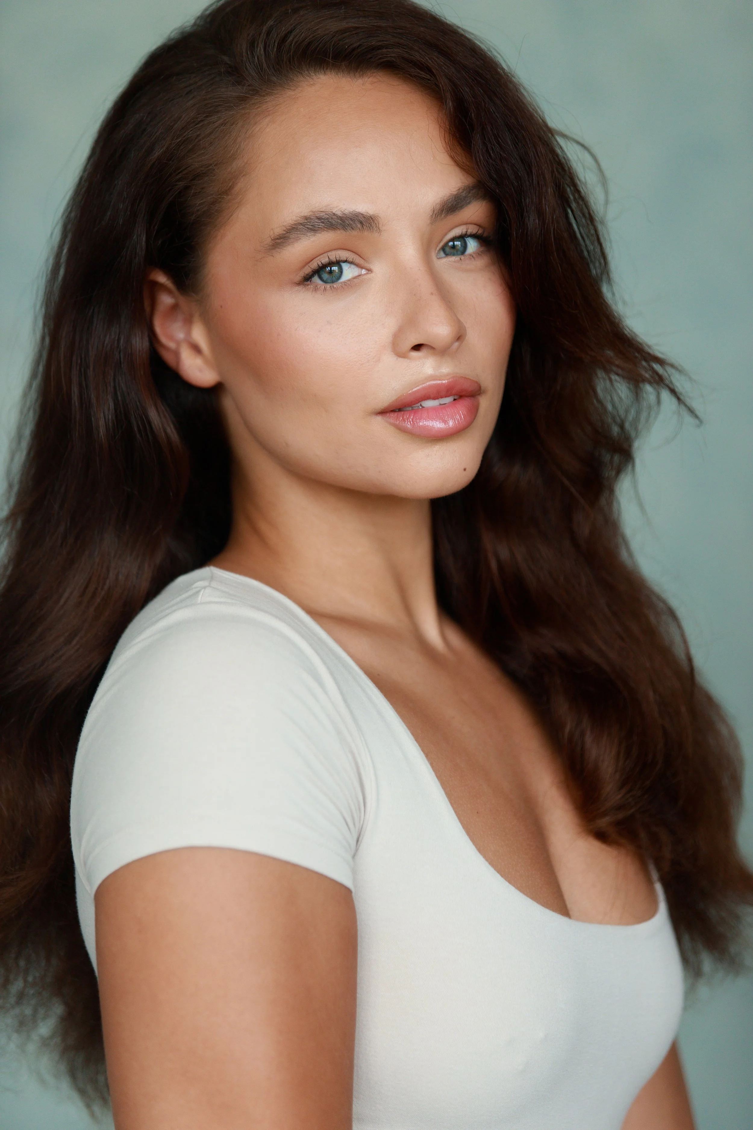 Portrait of a woman with long wavy brown hair and blue eyes, wearing a white top, looking at the camera with a neutral expression.
