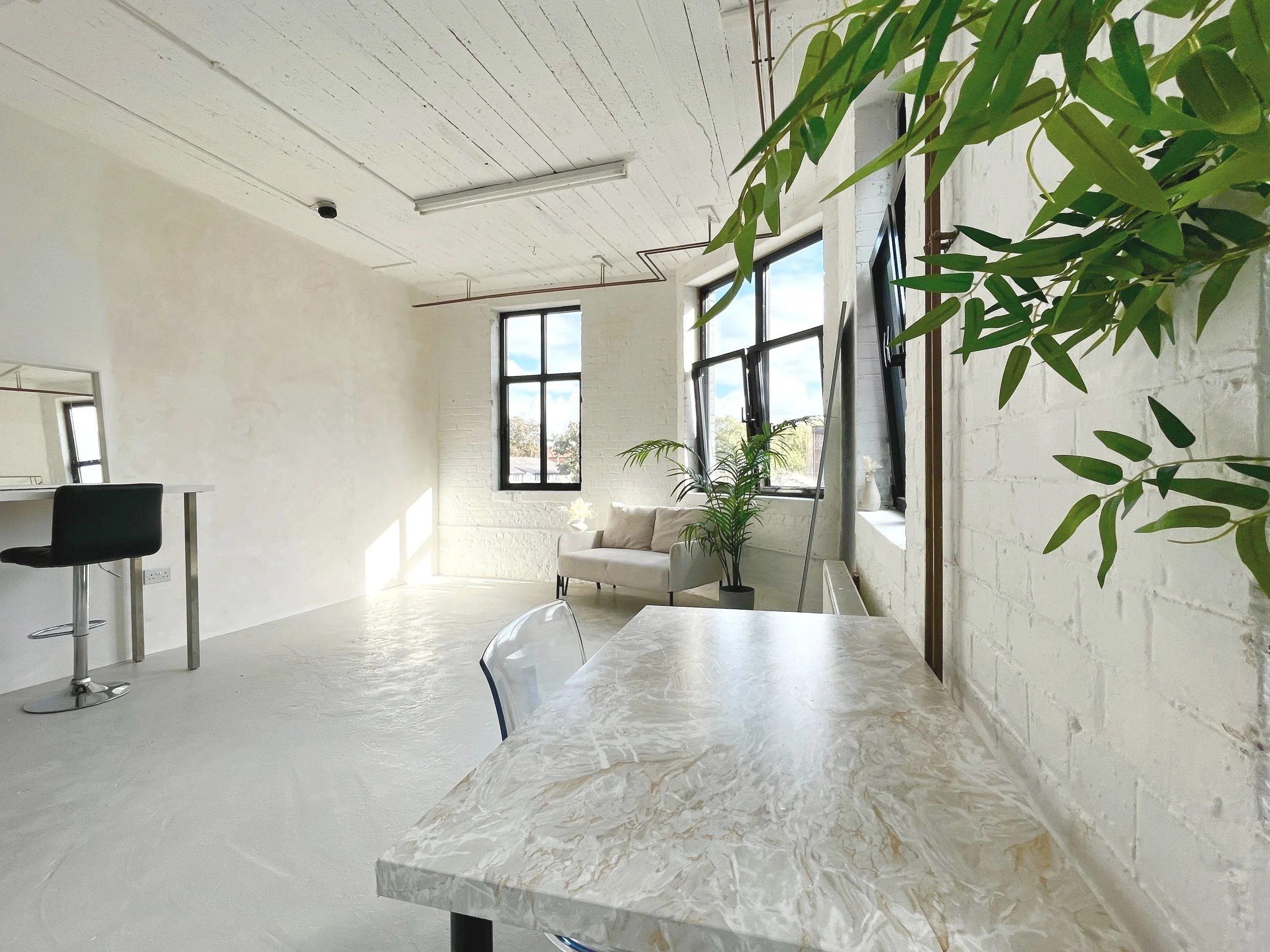 Bright, minimalistic living space with white brick walls, large windows, a beige sofa, a marble table, and green plants.