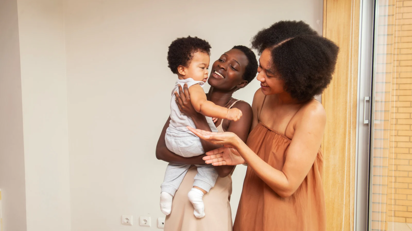 Two women smiling at a baby they are holding indoors.