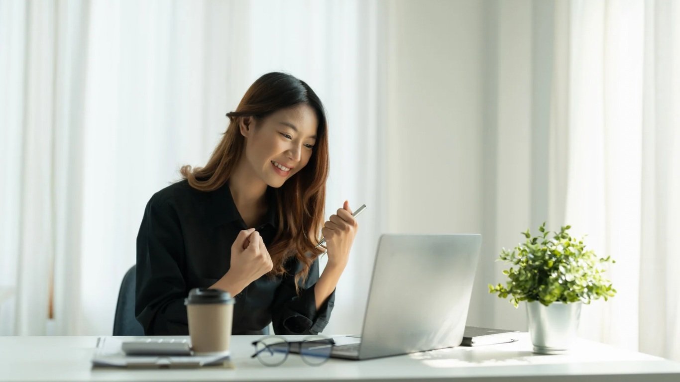 Woman smiling and sitting at a table with a laptop, coffee cup, glasses, and plant, appearing happy or excited.