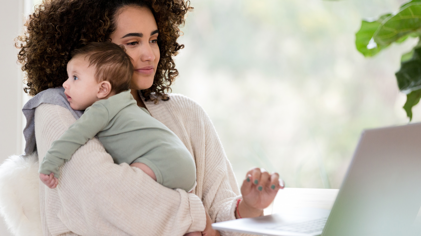 A woman holding a baby while working on a laptop at home.