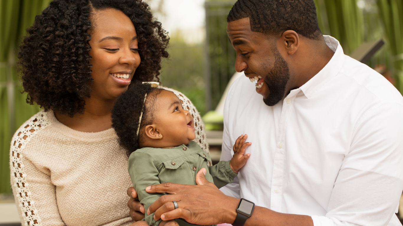 A happy family with parents holding a baby, all smiling and looking at each other.