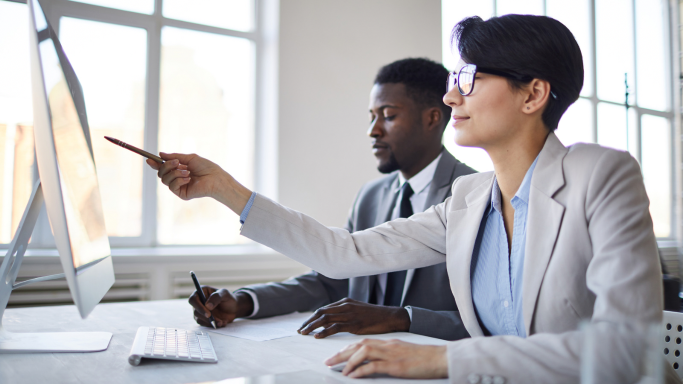 Two business professionals in suits working at a computer, woman pointing at screen with pencil, man writing notes.