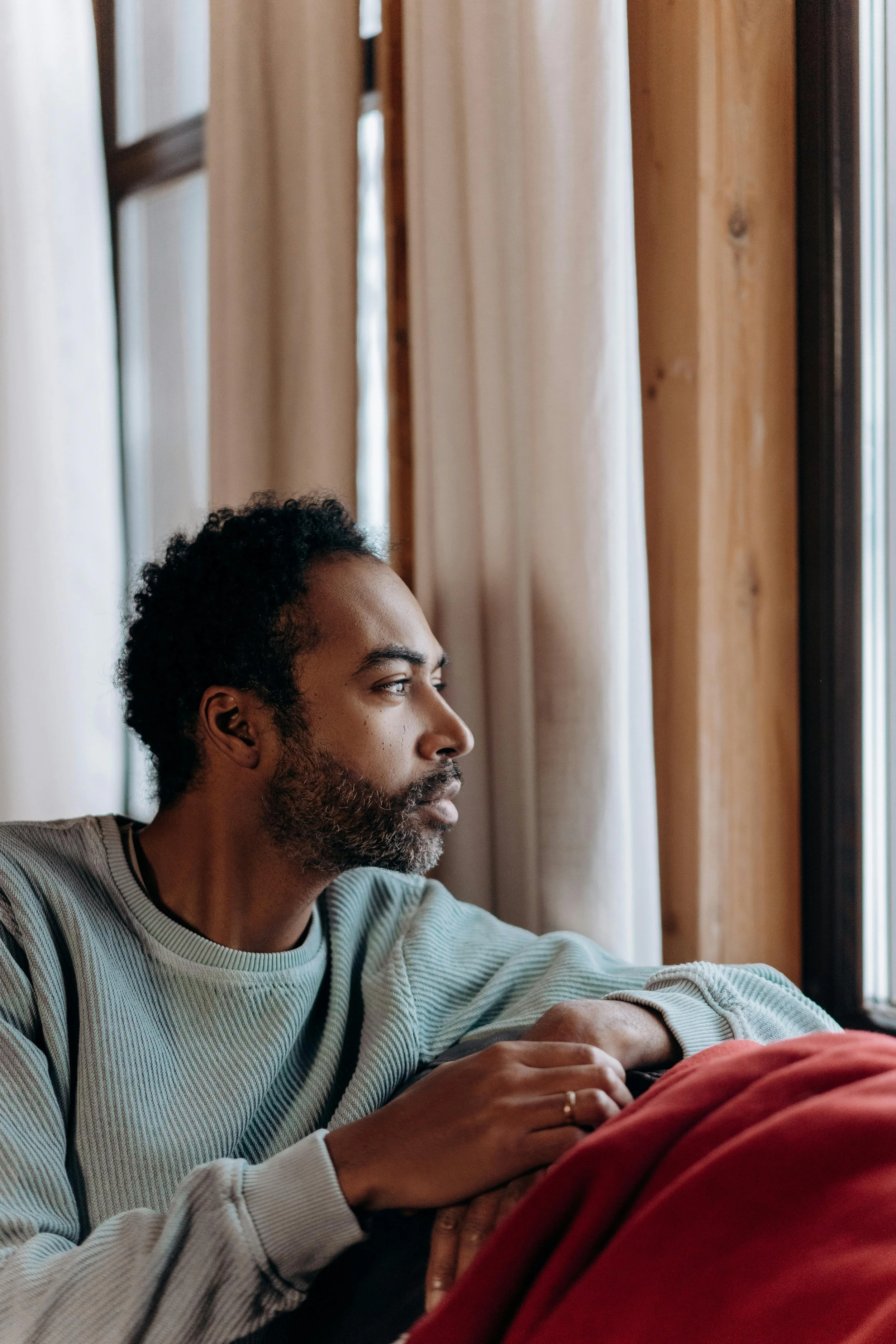 A Black man sitting quietly by a window in deep thought, reflecting on a difficult family distance