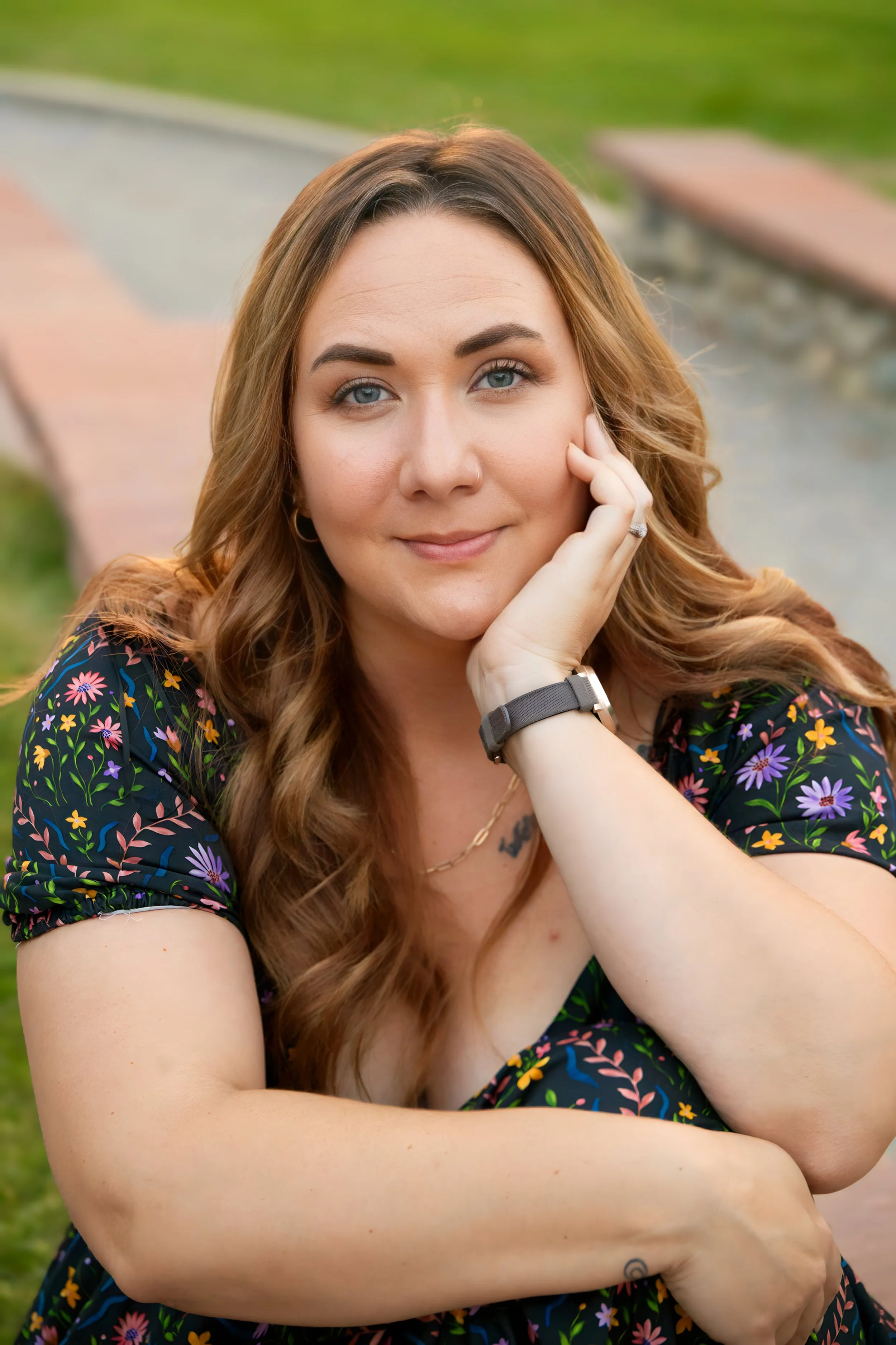 A woman with long, wavy, light brown hair, blue eyes, wearing a floral black dress, resting her face on her hand outdoors near a park bench and a curved pathway.