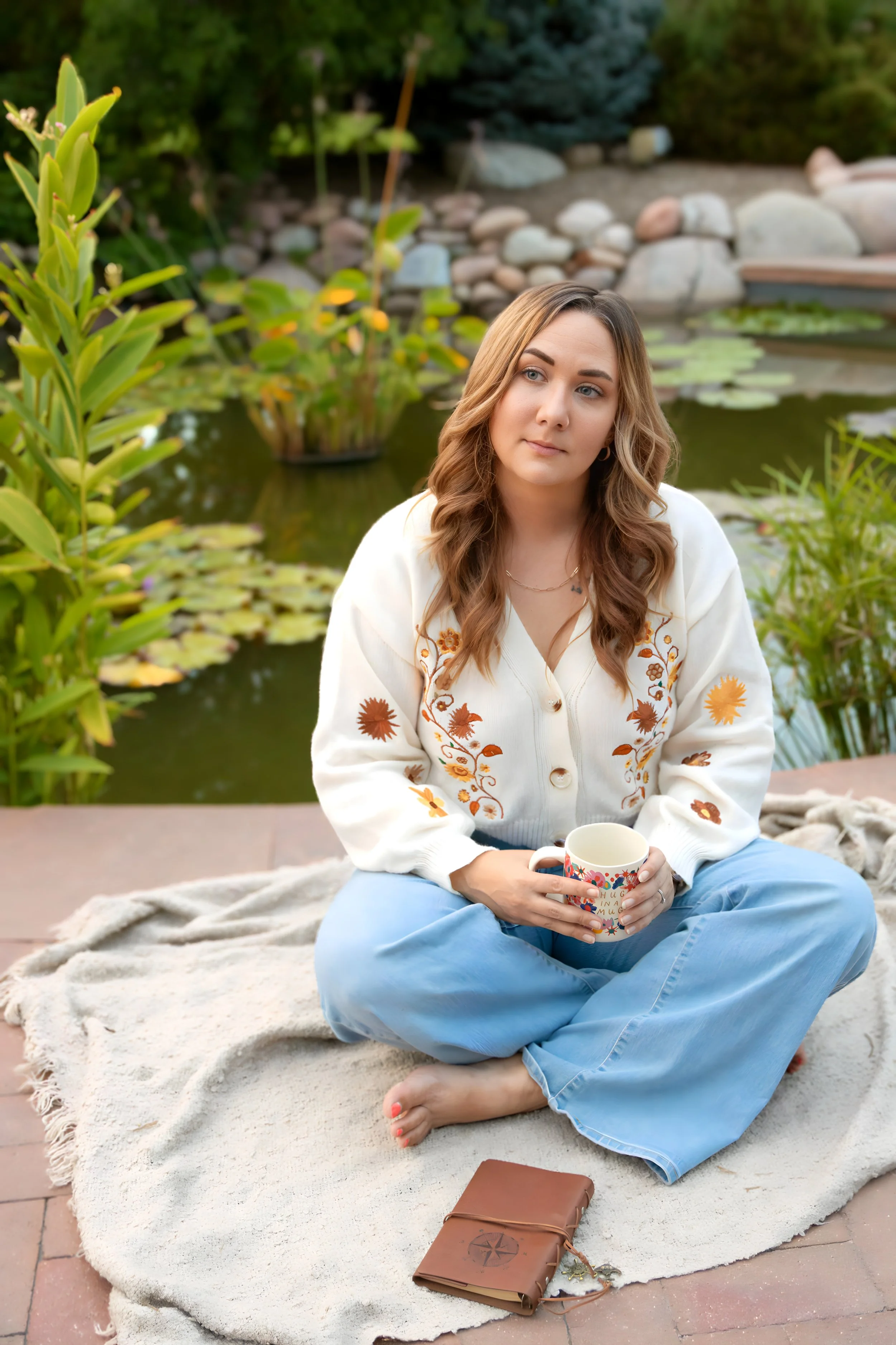 A woman with wavy blonde hair sitting cross-legged on a blanket beside a pond. She is holding a colorful mug, wearing a white embroidered cardigan and blue jeans. There is a brown leather journal with a compass design on the ground in front of her.