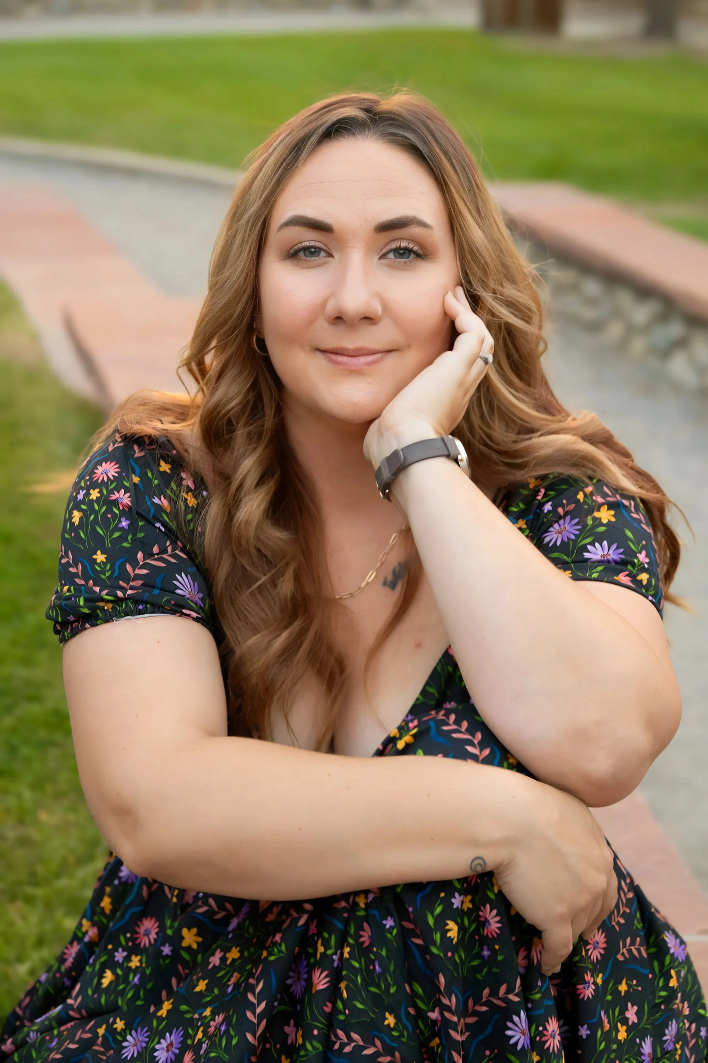 A woman with long, wavy, light brown hair, wearing a black dress with a colorful floral pattern, sitting outdoors on a park bench.