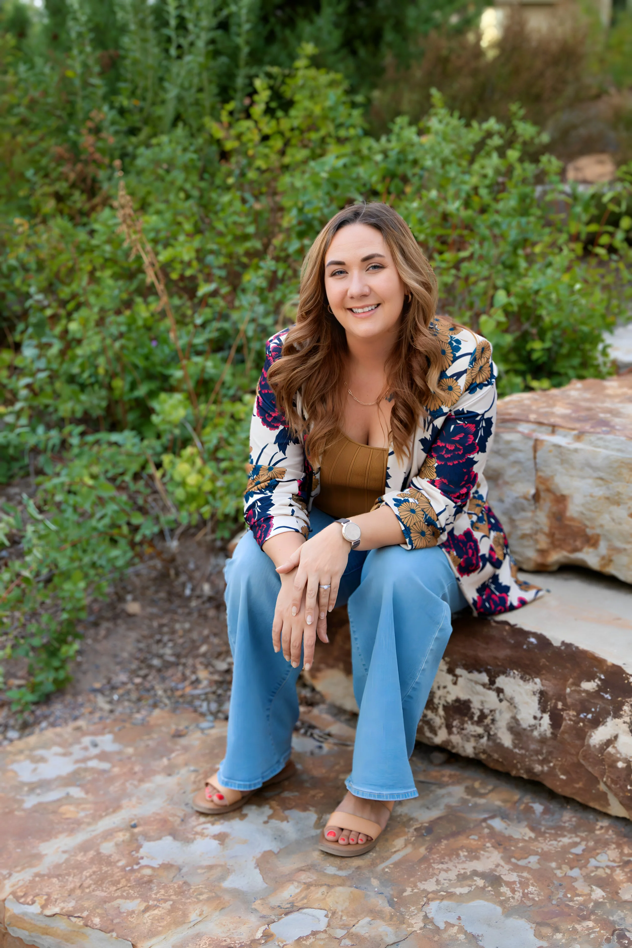 A woman sitting on a rock outdoors surrounded by green foliage, smiling at the camera. She has long wavy hair, is wearing a floral blazer, a brown top, blue jeans, and sandals.