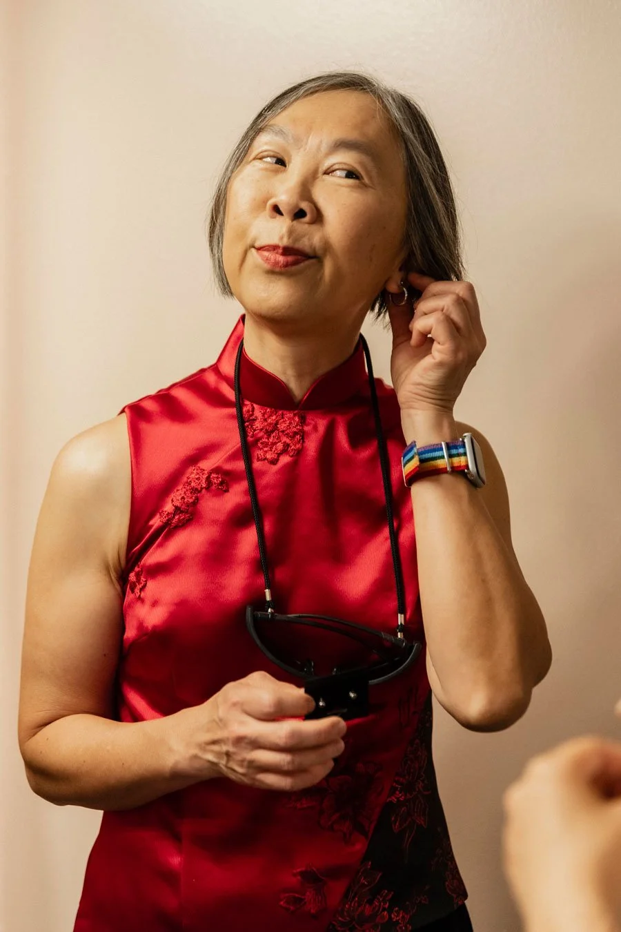 A woman with short gray hair wearing a red traditional Chinese sleeveless dress, holding glasses around her neck, and adjusting her earring.