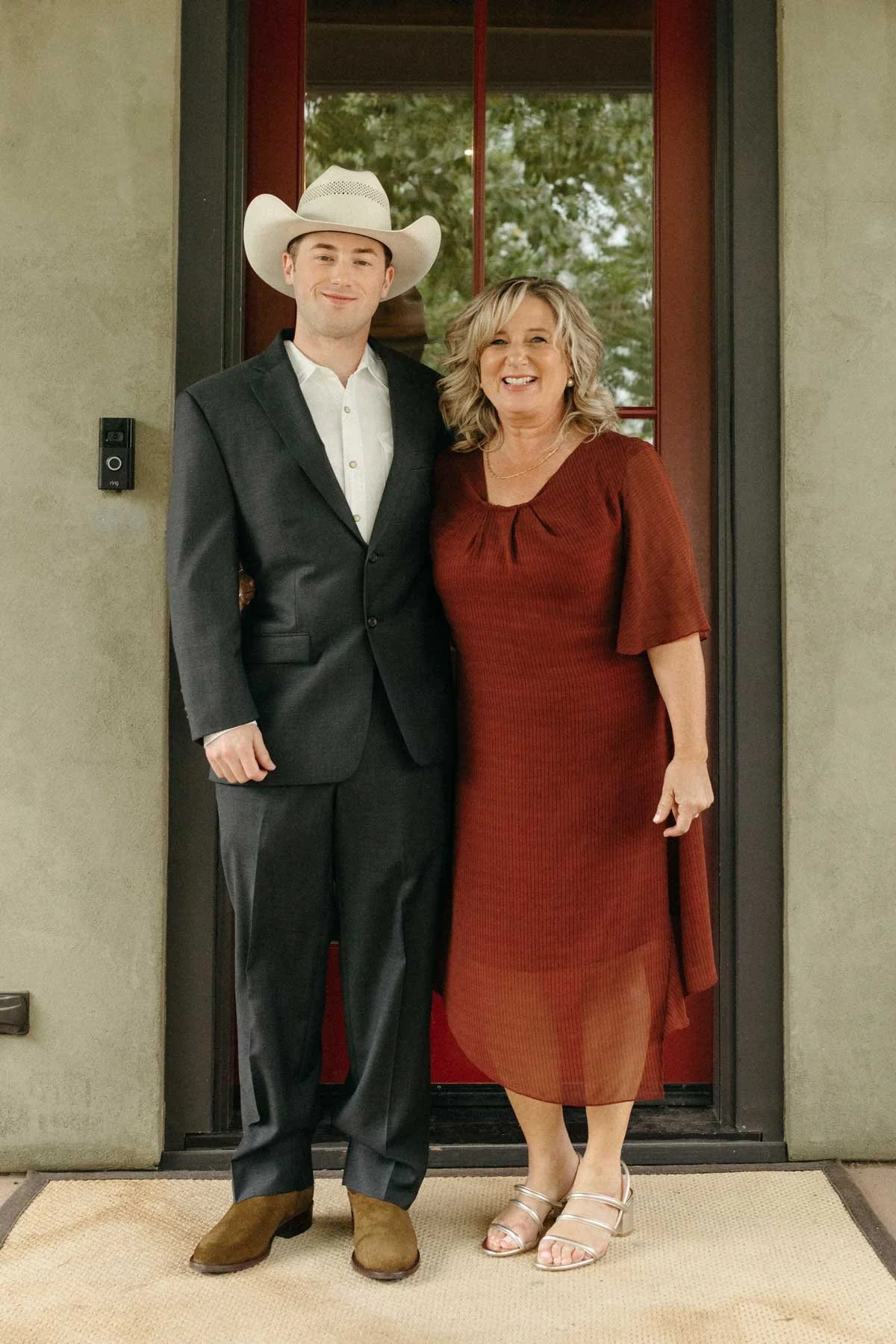 A young man in a suit and cowboy hat standing next to a woman in a red dress, both smiling, in front of a doorway.