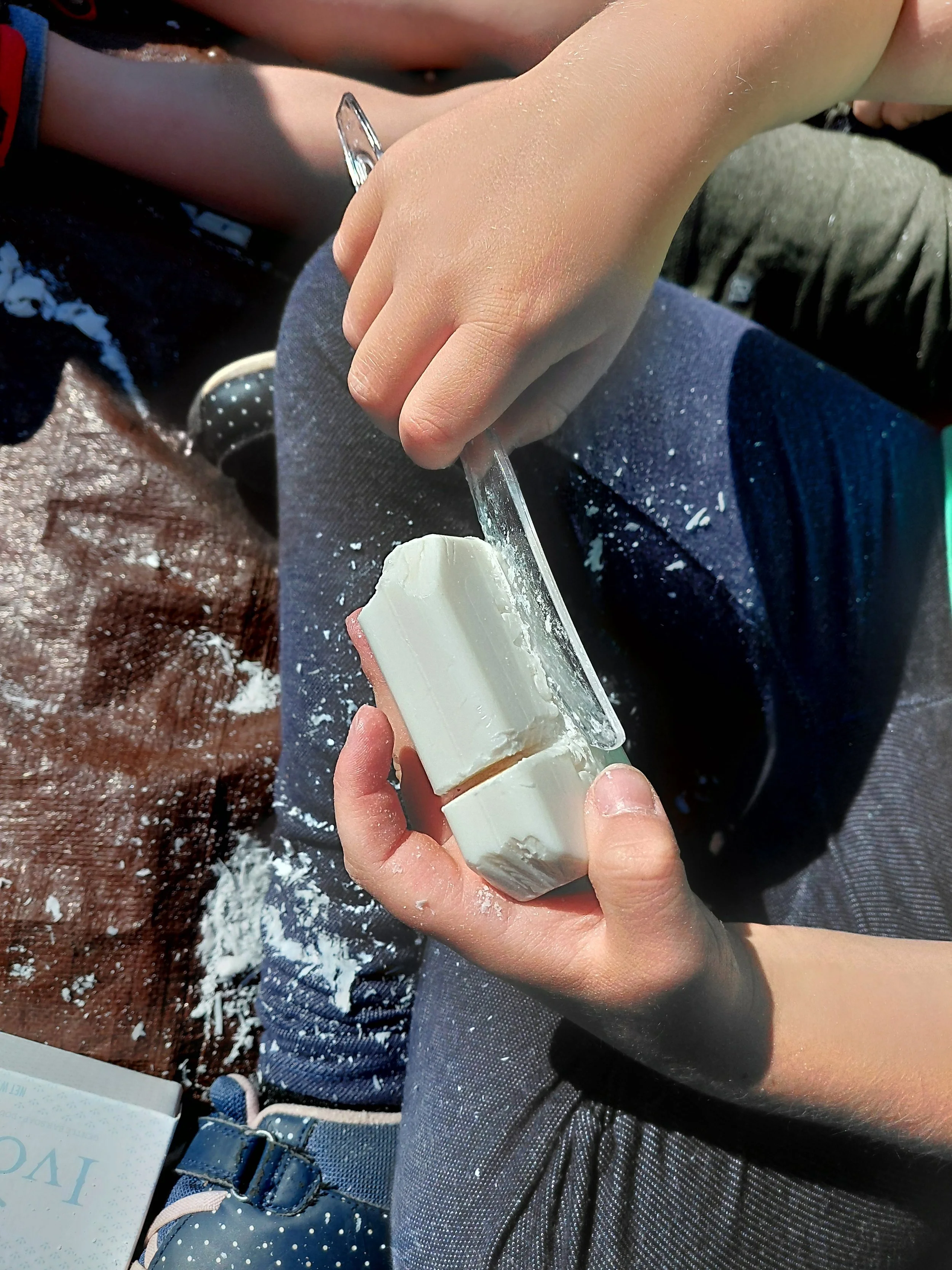 Child's hand with a small knife scraping white wax from a block of wax, sitting on a surface with wax shavings scattered around.
