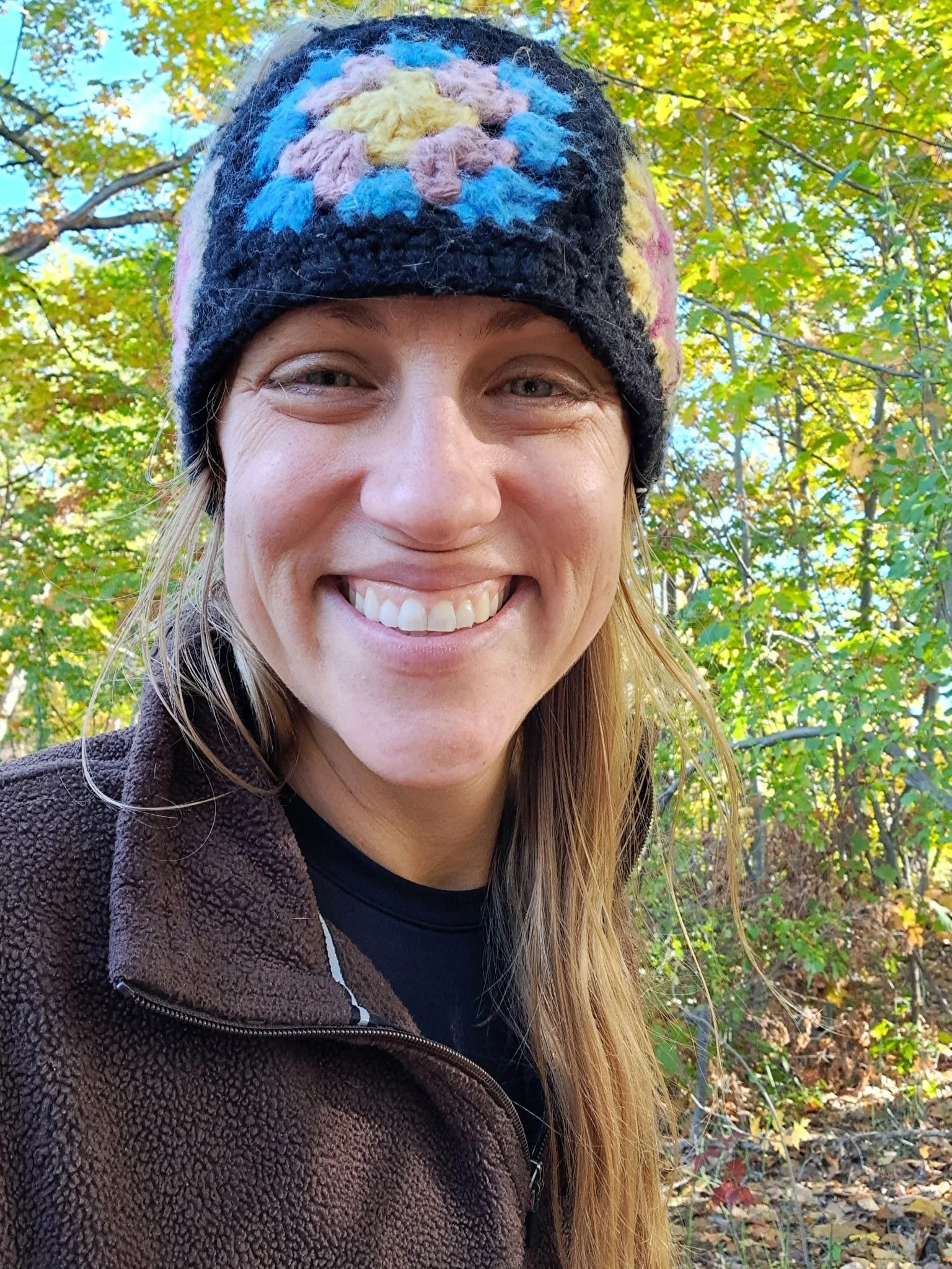 A woman smiling outdoors in autumn, wearing a colorful knit hat and a brown fleece jacket, with fall foliage in the background.