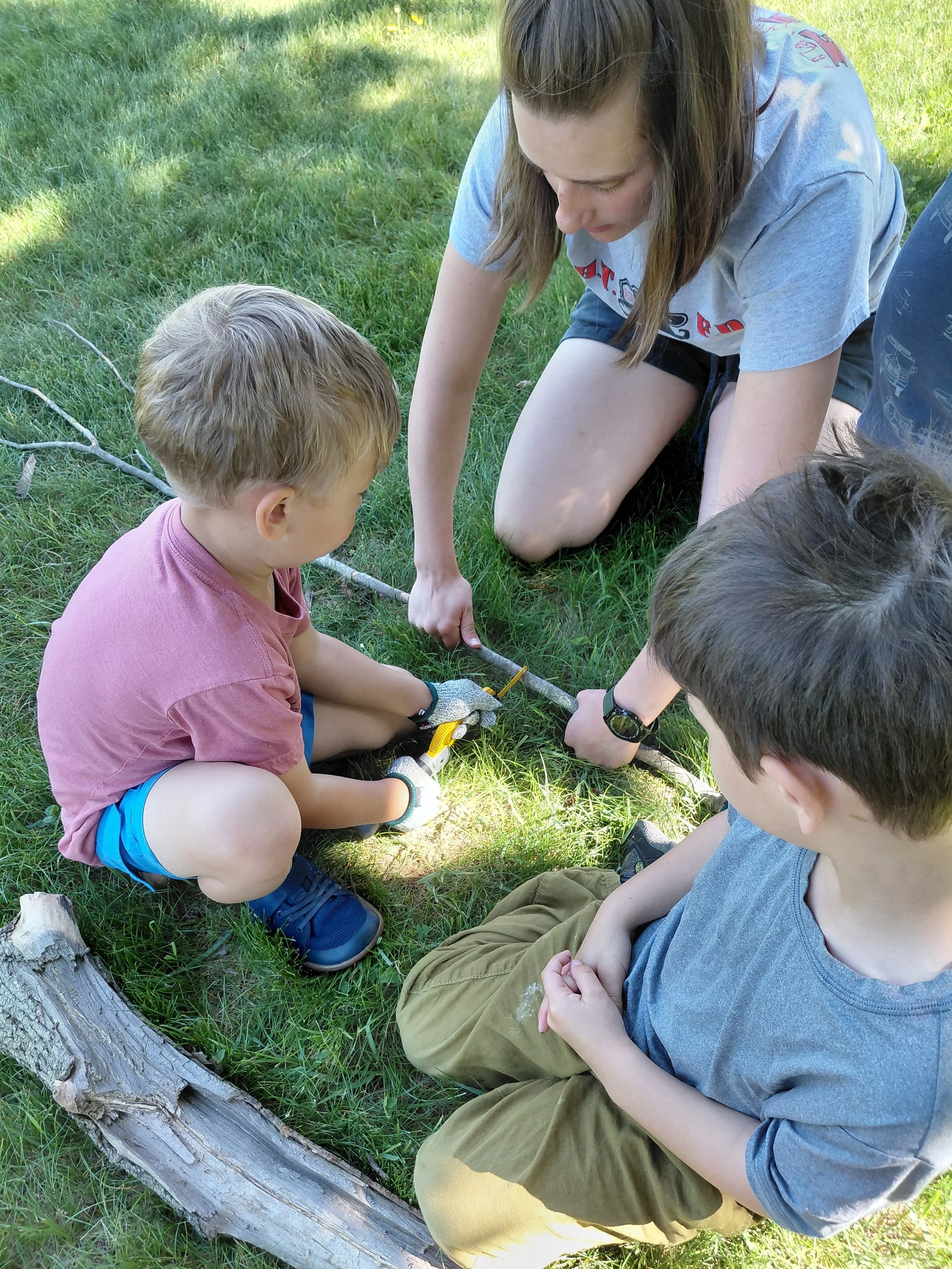 Three children and one young woman kneeling on grass outdoors, examining something on the ground near a fallen tree.