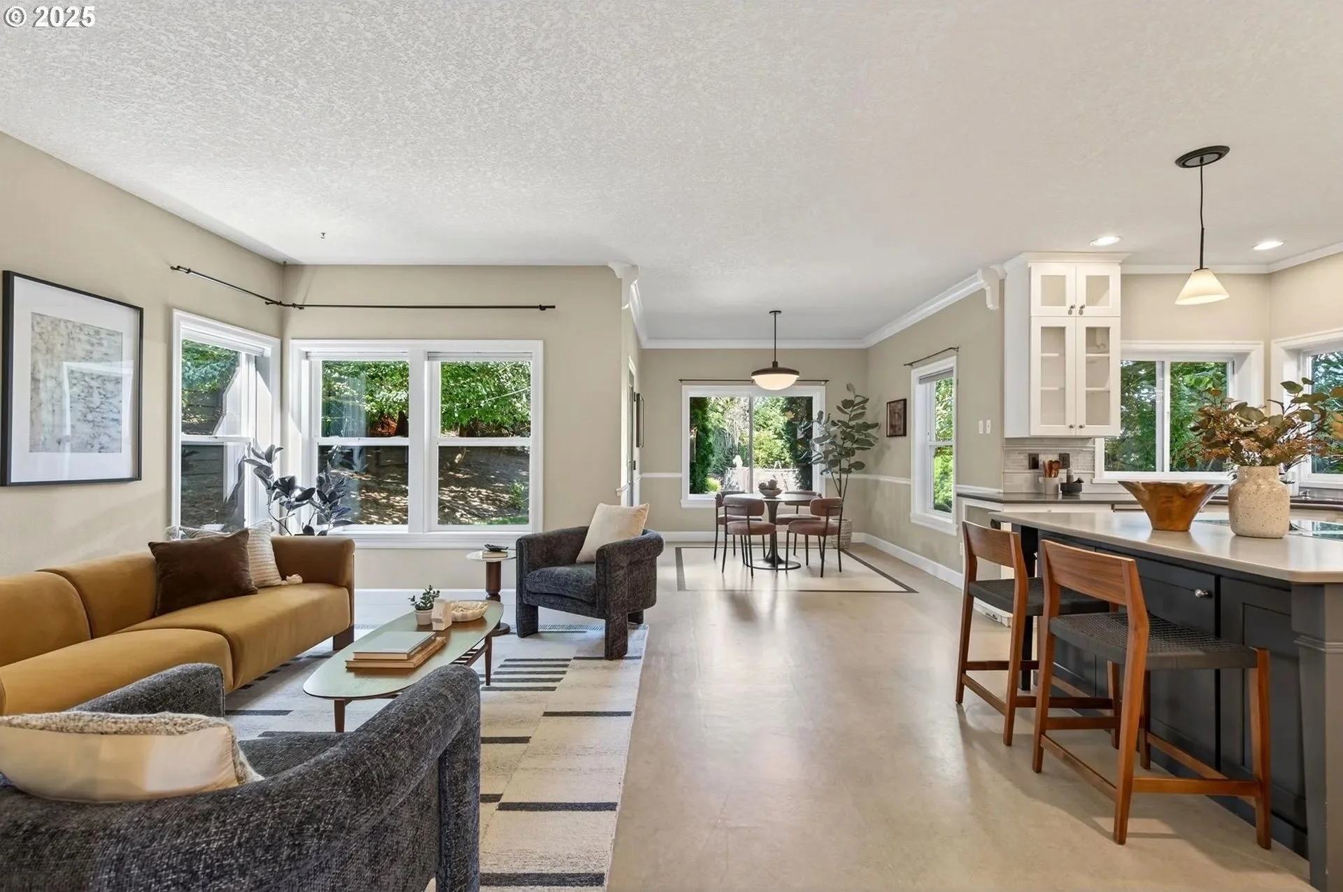 Open living room and kitchen with large windows and natural light, beige walls, and hardwood flooring. The living room has a mustard yellow sofa, dark gray armchair, coffee table, and framed artwork on the wall. The kitchen features a white counterto