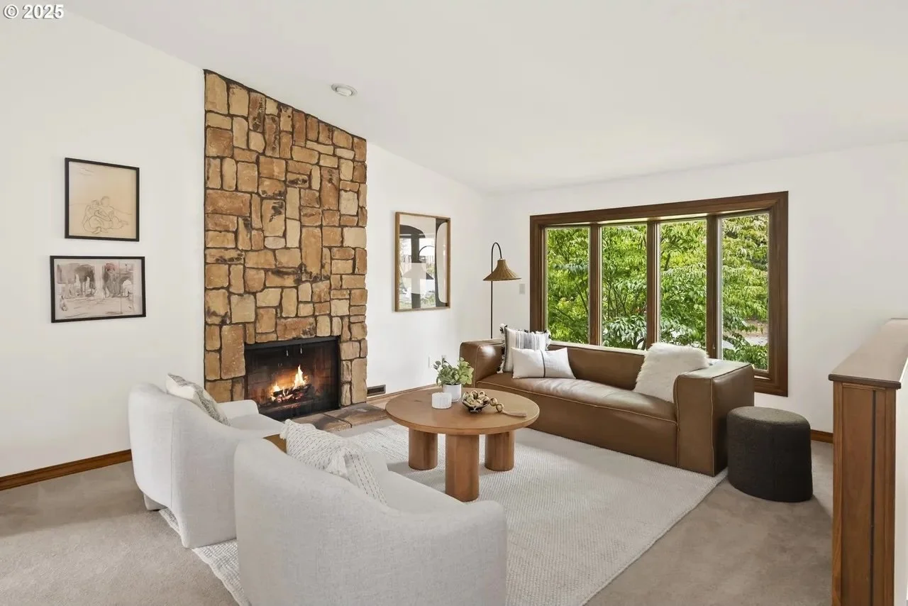 Living room with a stone fireplace, a brown leather sofa, a white armchair, large windows with greenery outside, and a round wooden coffee table.
