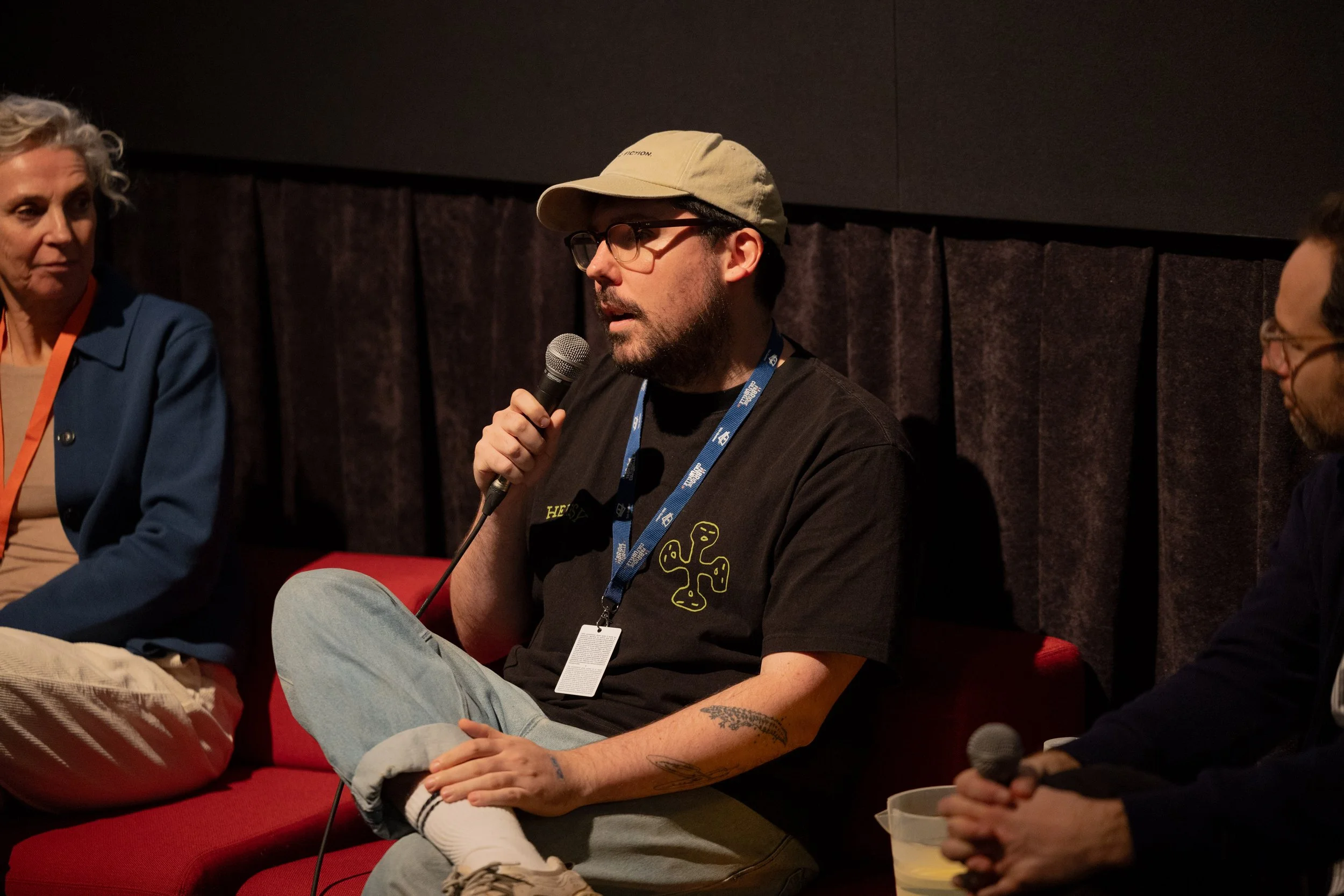 Man speaking into microphone during panel discussion, sitting on red chair, wearing glasses, beige cap, black T-shirt with yellow graphic, and light jeans with tattoos on his arms.