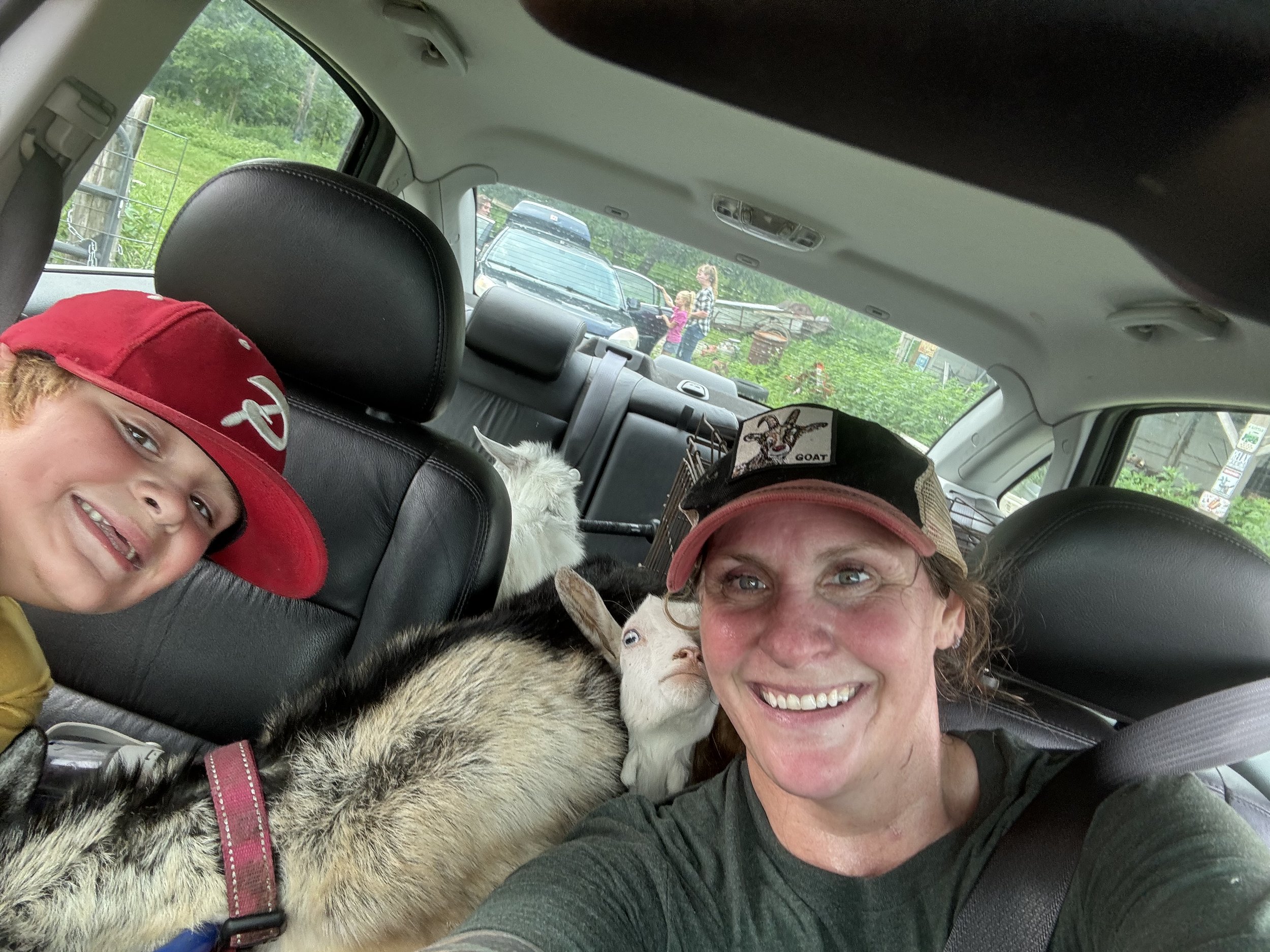 A young boy wearing a red baseball cap smiling, sitting between an older woman with curly hair and a white and gray dog in the backseat of a car. Two people are visible outside through the rear window, a woman with curly hair and a child standing nea