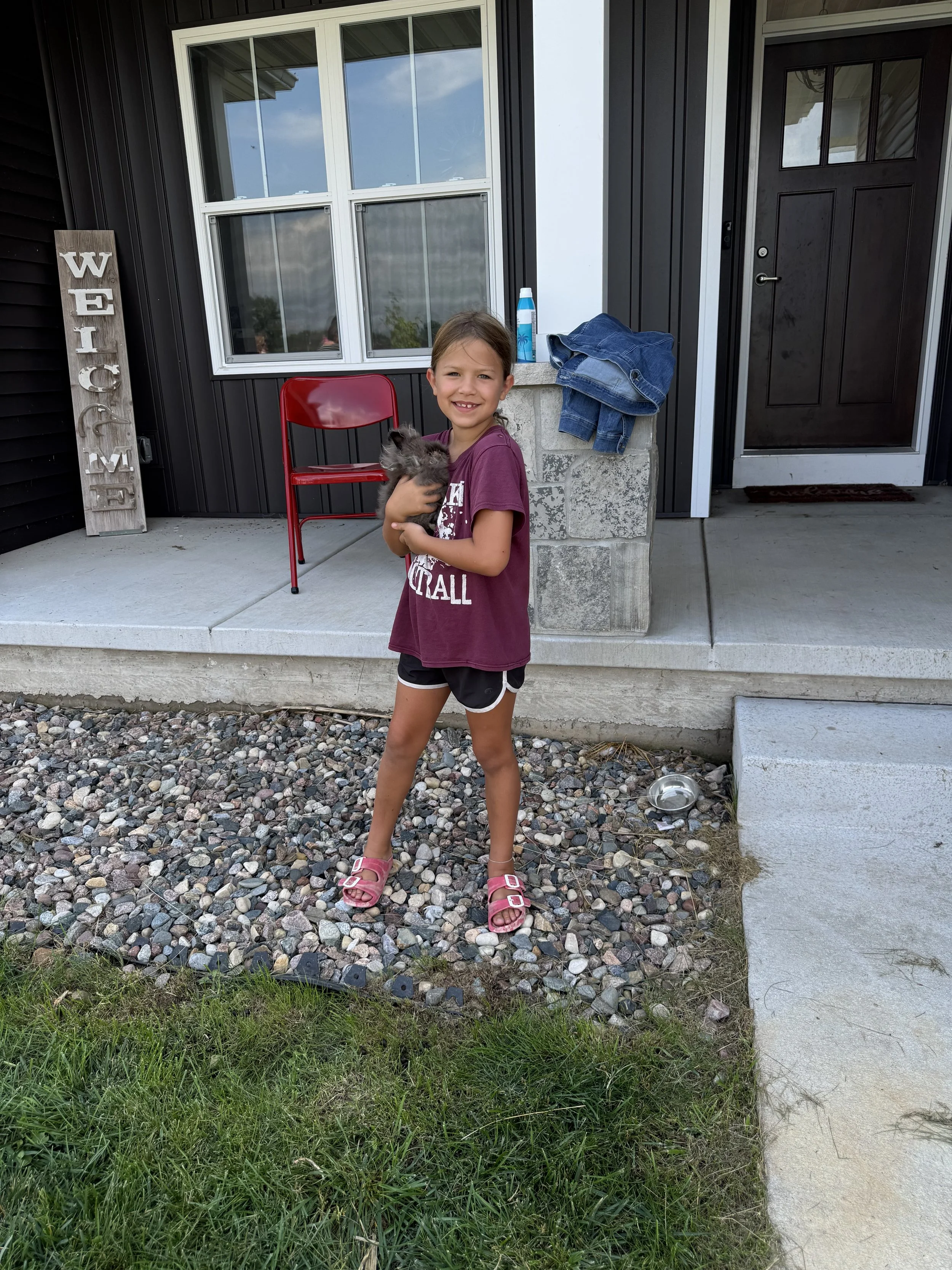 A young girl with brown hair, wearing a maroon t-shirt, black shorts, and pink sandals, stands outside a house, holding a fluffy gray kitten. The house has black siding, a white window, a dark door with glass, a gray concrete porch, and a welcome sig