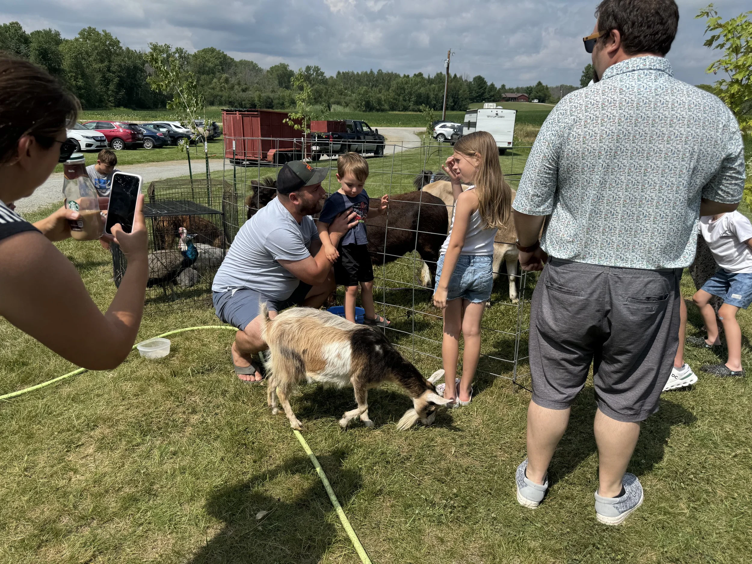 Group of people interacting with goats and a small farm animal pen outdoors on a sunny day with a grassy field and parked cars in the background.