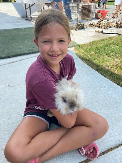 A young girl sitting on the ground outdoors, smiling and holding a fluffy white rabbit.