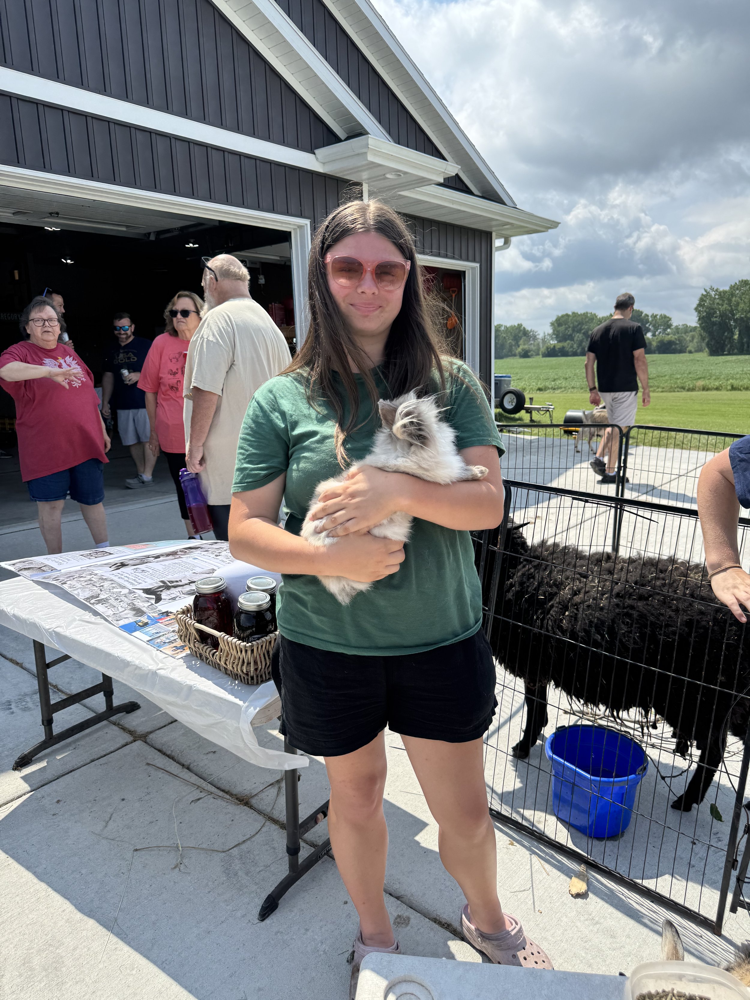A young woman in pink sunglasses is holding a small fluffy white and gray kitten outside near a cage. The background includes a group of people near an open garage, a table with jars and papers, and a blue bucket. The setting appears to be a backyard