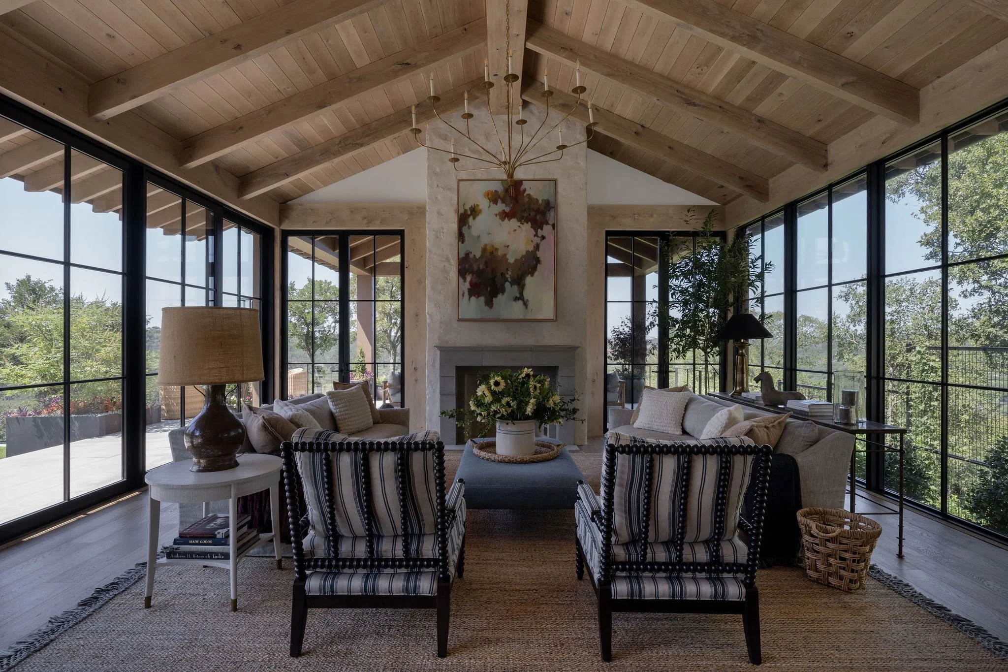 Living room with large floor to ceiling glass windows, wooden vaulted ceiling, sitting area with sofas and striped armchairs, a central coffee table with a flower arrangement, a fireplace, and artwork on the mantel.