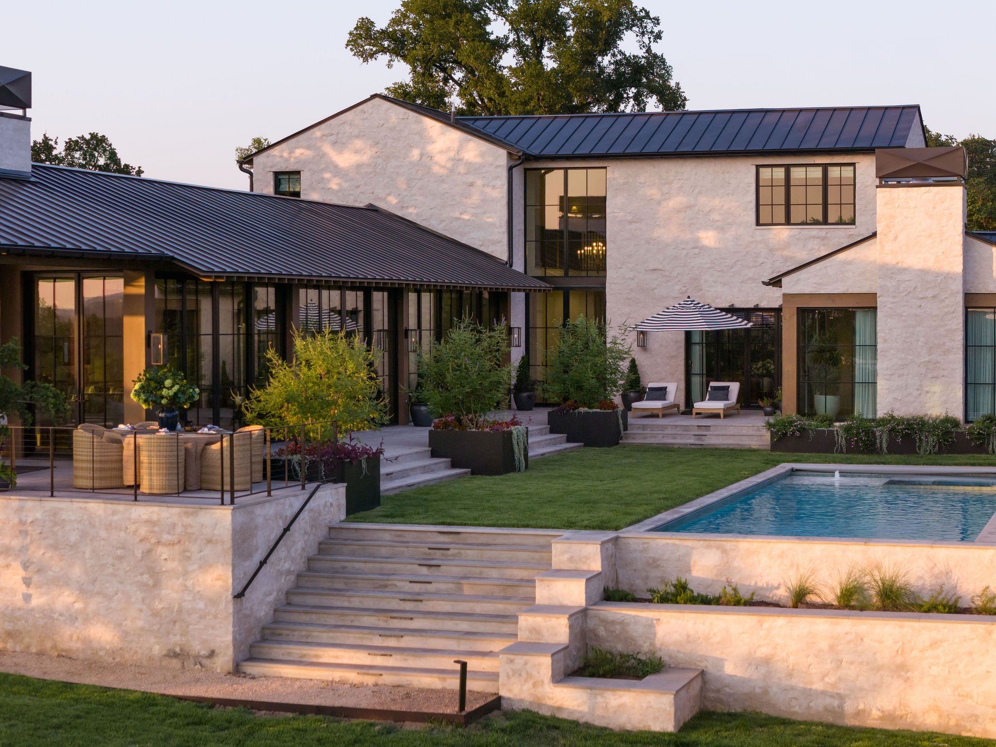 Beautiful modern backyard with a custom swimming pool, outdoor seating area with chairs and table, lawn, and a multi-story house with large custom surround windows and a metal roof, during sunset.