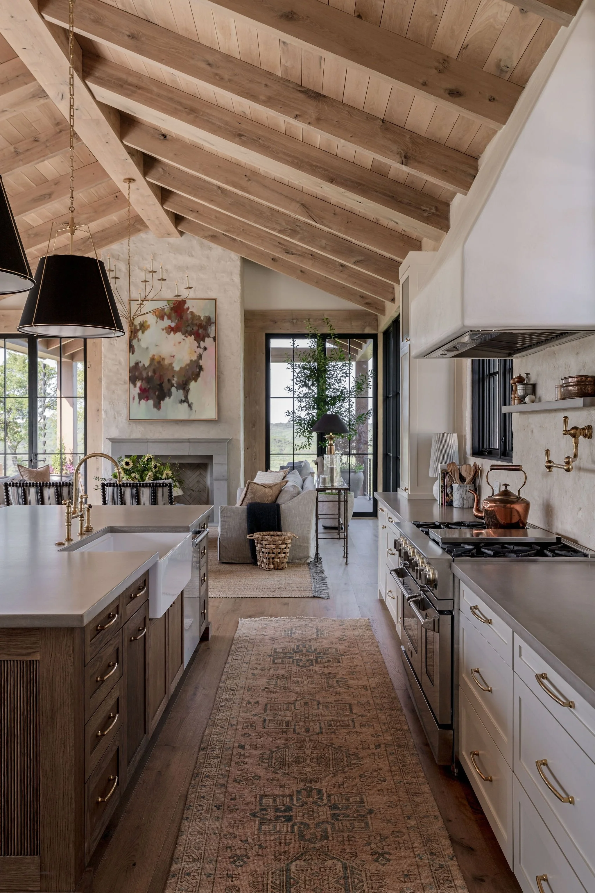 Open-concept custom kitchen and living room with rustic wood white walnut ceiling, modern white and wood cabinets, top of the line stainless steel appliances, black pendant lights, patterned rug, and a sitting area with sofa near large windows.