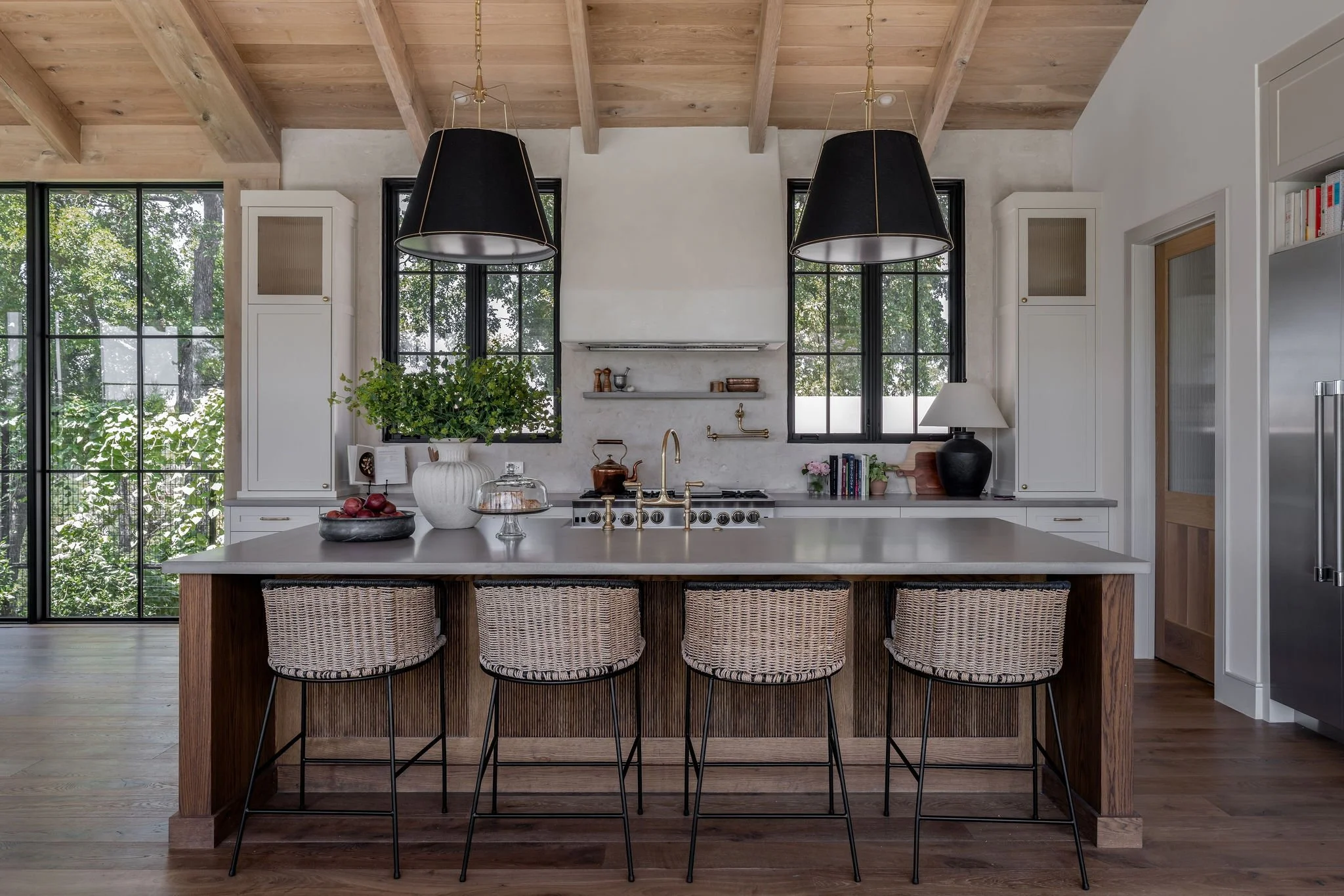 Modern kitchen with a large island, black pendant lights, white cabinets, and black-framed windows showing greenery outside.