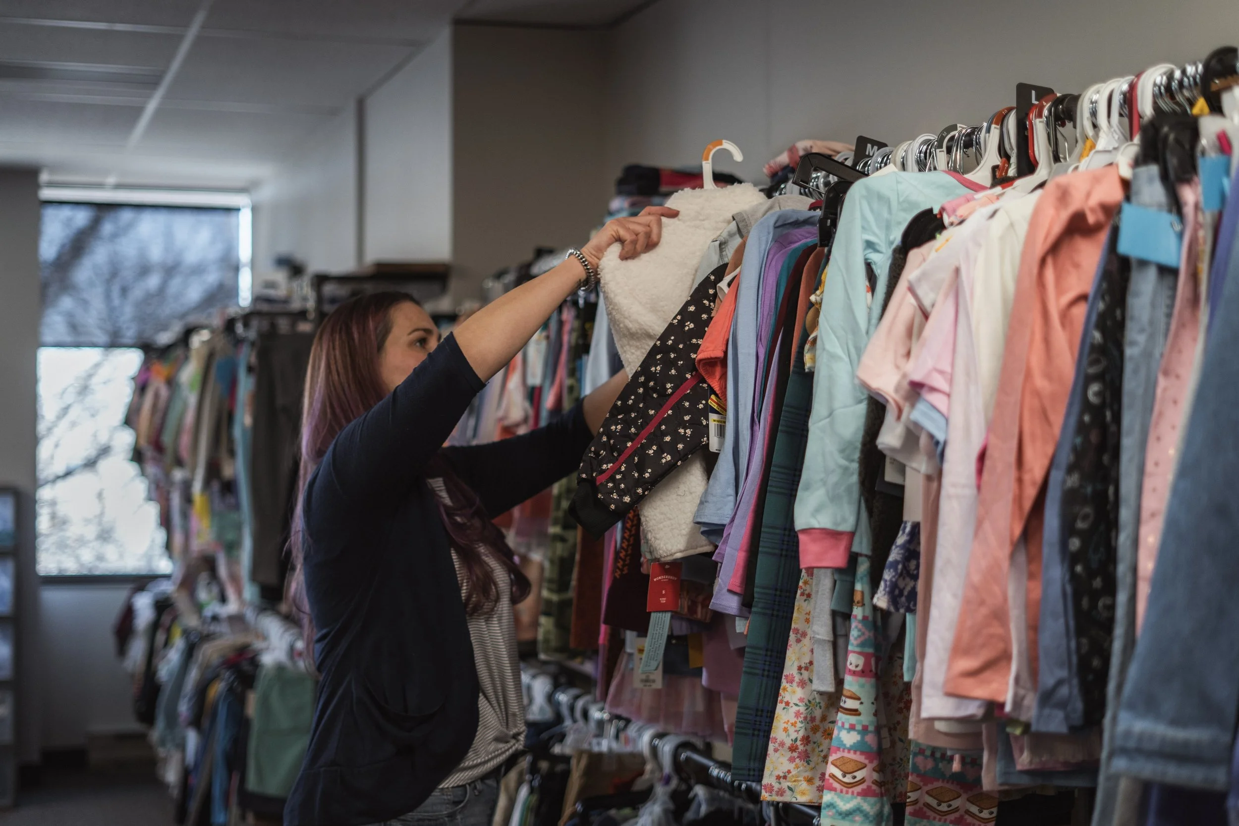 A woman is browsing clothes on a rack in a foster closet. She is reaching for a piece of clothing and the rack is filled with various colorful outfits.