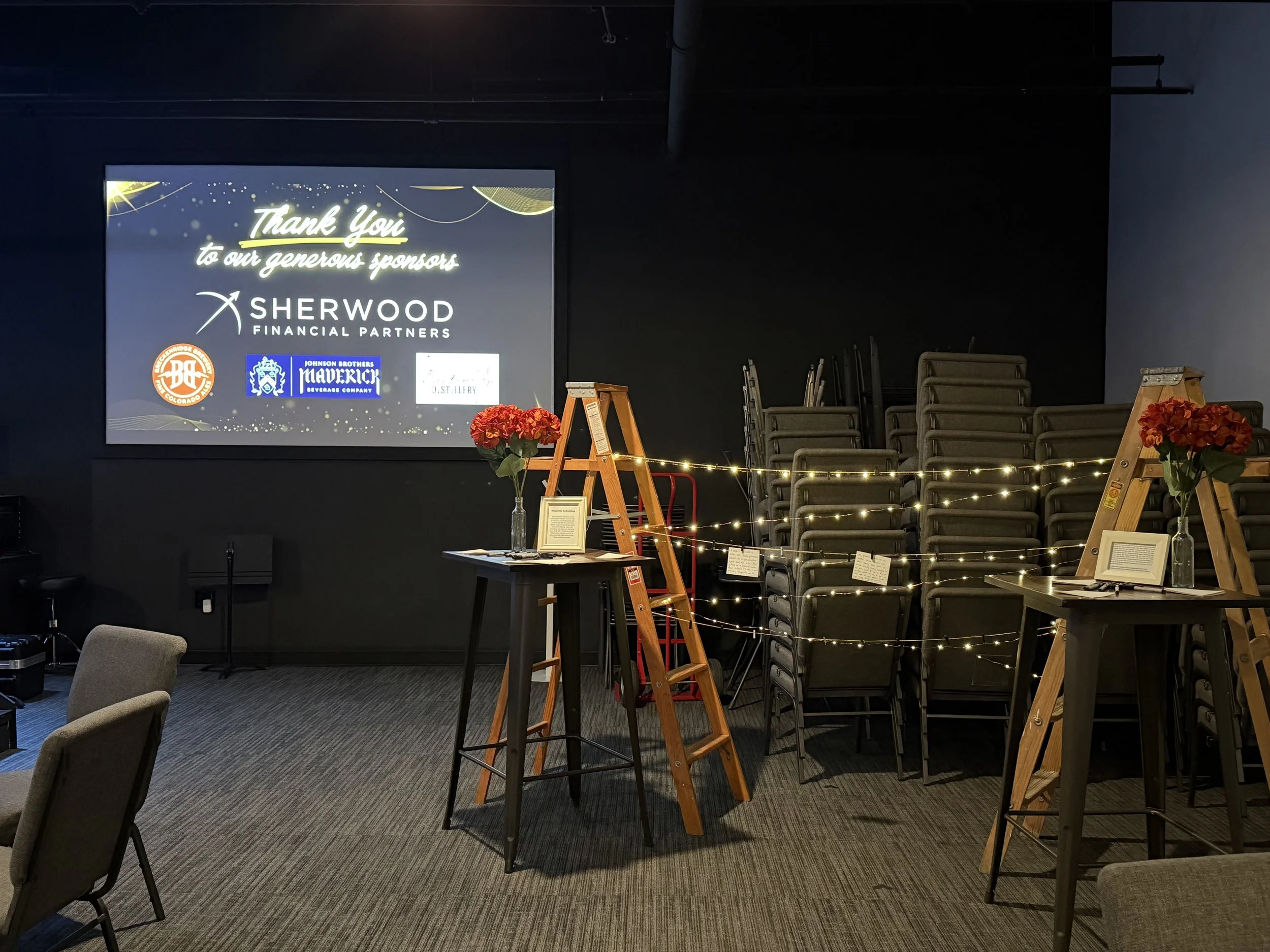 A presentation screen displaying a thank you message and logos, surrounded by chairs and flower arrangements in a dimly lit room.