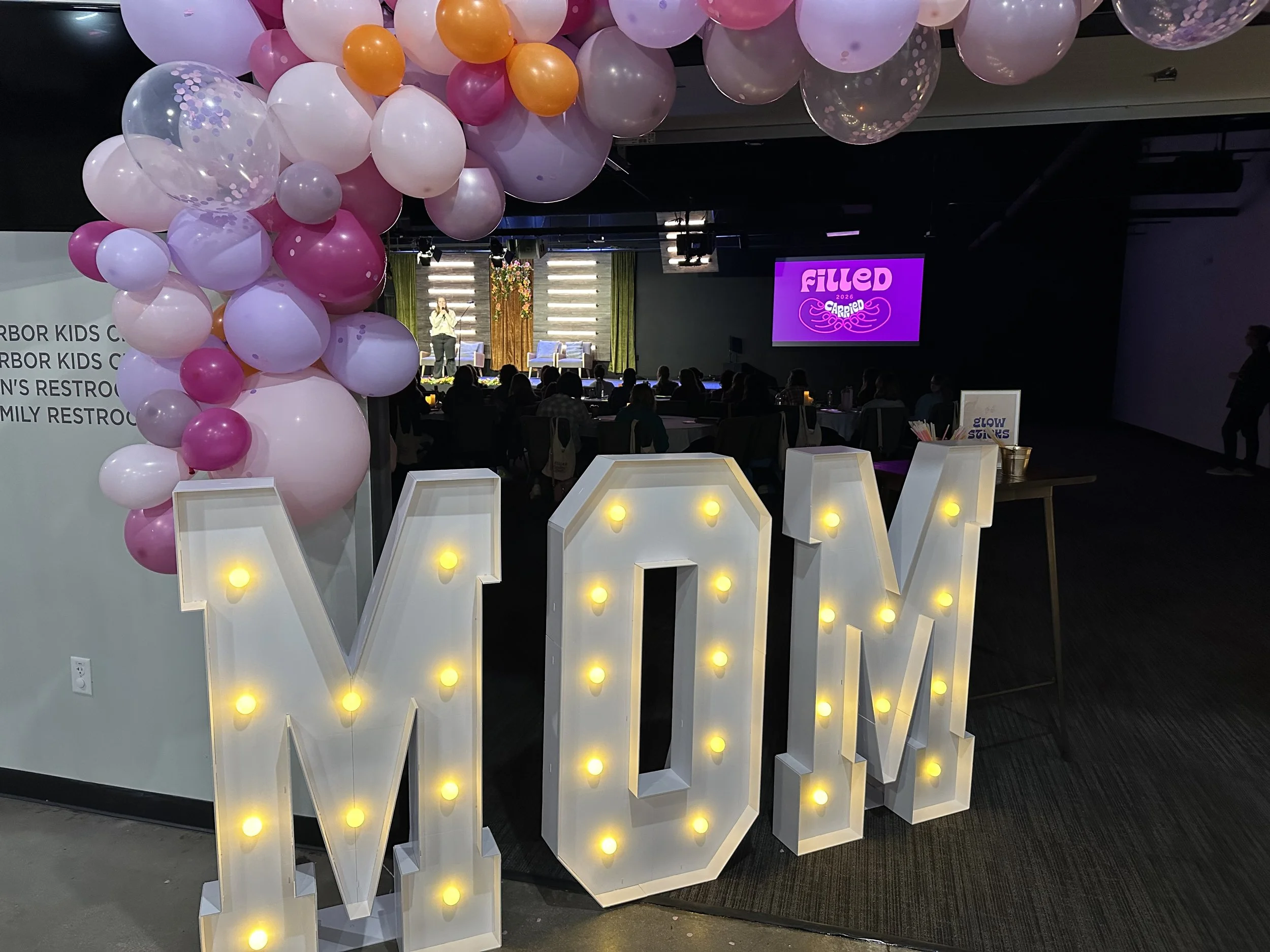 Pink and purple text on a beige background reads "Filled Local Denver 2026" with a heart-shaped design made of two hands holding a word "Carried". Additional text states "A gathering for foster, adoptive, & kinship moms, May 1 - May 2, Denver, CO."