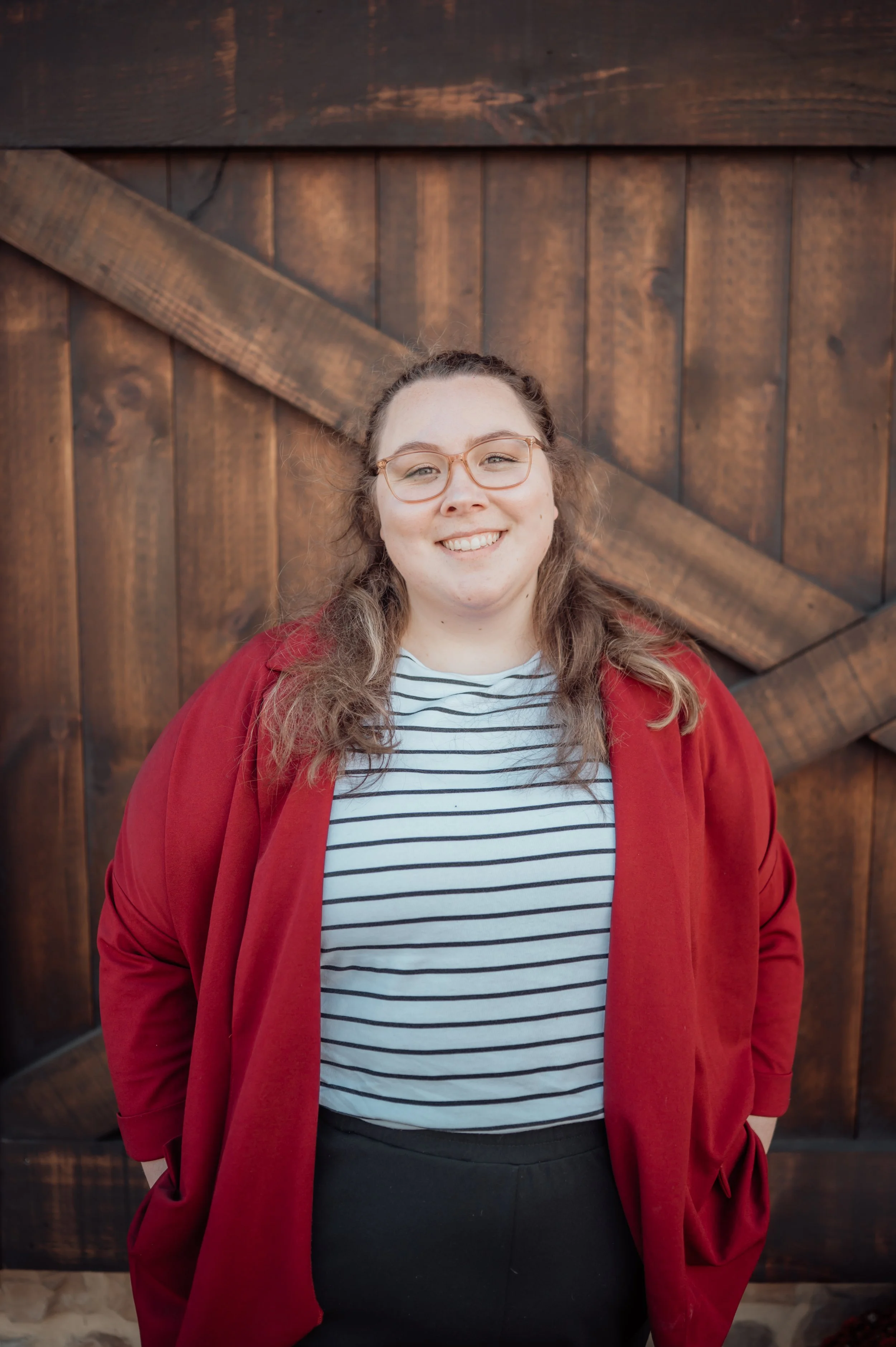 A smiling woman standing outdoors in front of a wooden barn door, wearing glasses, a striped t-shirt, and a red jacket.
