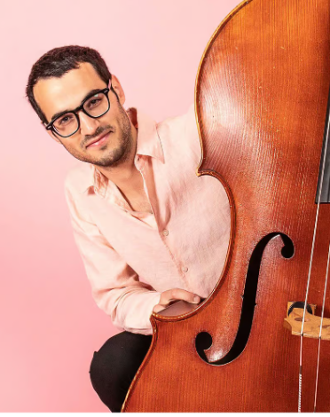 Smiling man wearing glasses and a pink shirt posing behind a large wooden upright bass against a pink background