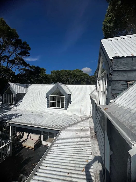 View of several white metal-roofed buildings with dormer windows, surrounded by trees under a clear blue sky.