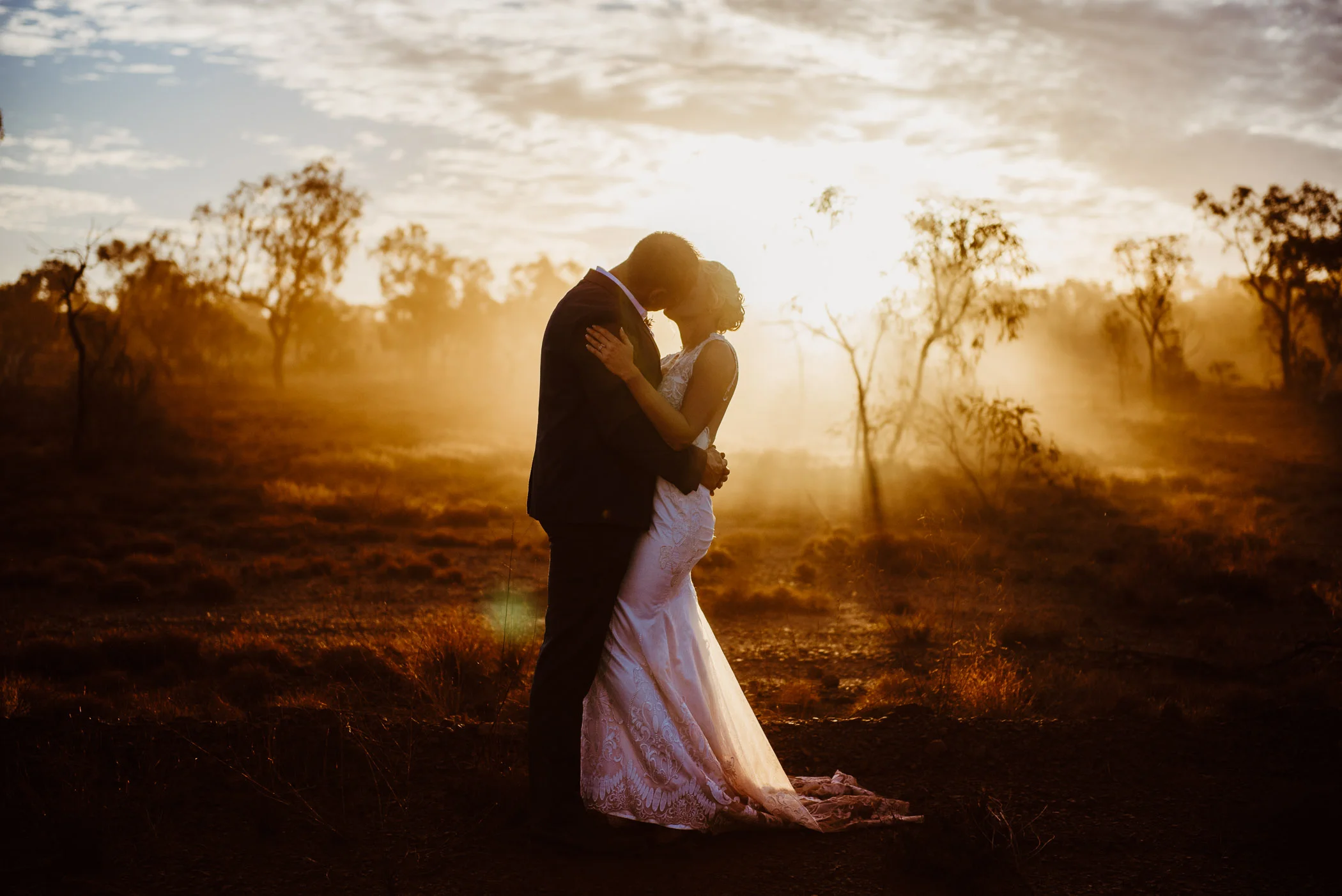 Couple kissing with darkened sunset shining over the red dirt outback landscape behind them on their wedding day