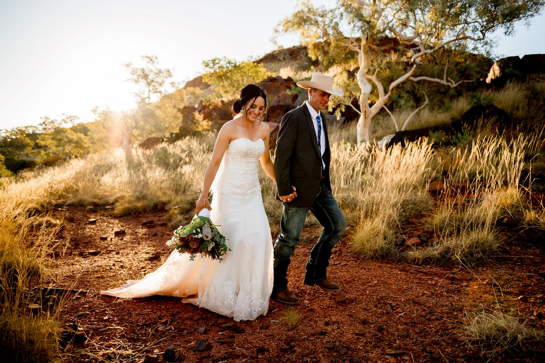 Bush Wedding couple walking hand in hand away from golden glow of the sun setting over the red hills behind them