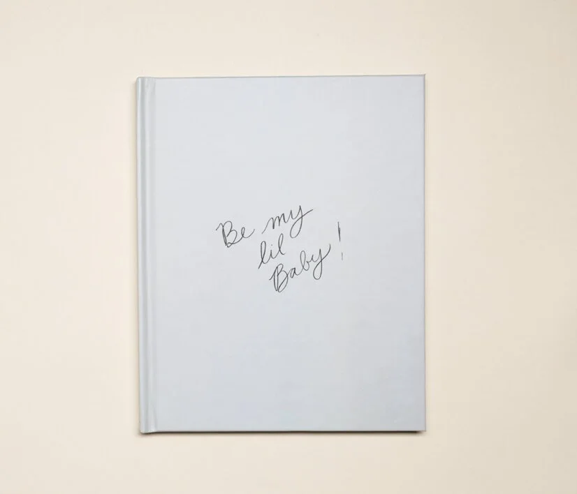 A white notebook with the handwritten message 'Be my lil Baby!' on the cover, placed on a plain beige background.