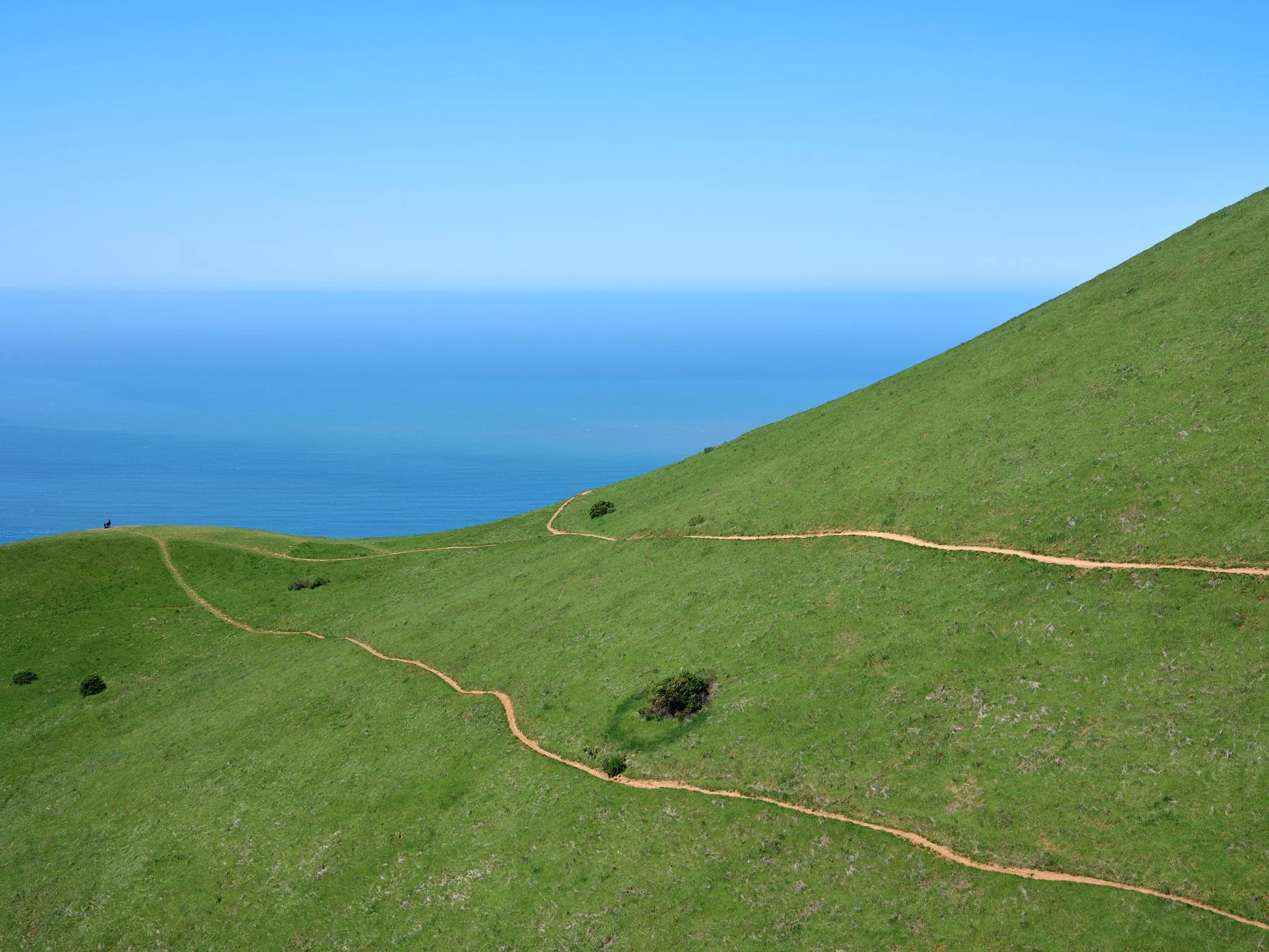 A lone figure sits on a A lone figure sits on a distant ridge overlooking the sea, framed by the descending lines of a coastal trail. This vast landscape emphasizes the feeling of being a 'perpetual observer' and reflects the distance I often feel na