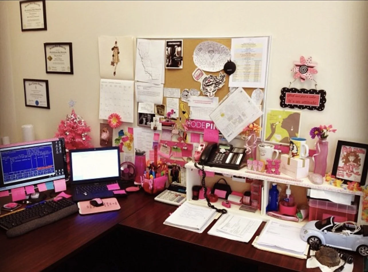 Cluttered office desk with pink decorations, computers, phones, and a bulletin board on the wall behind it.