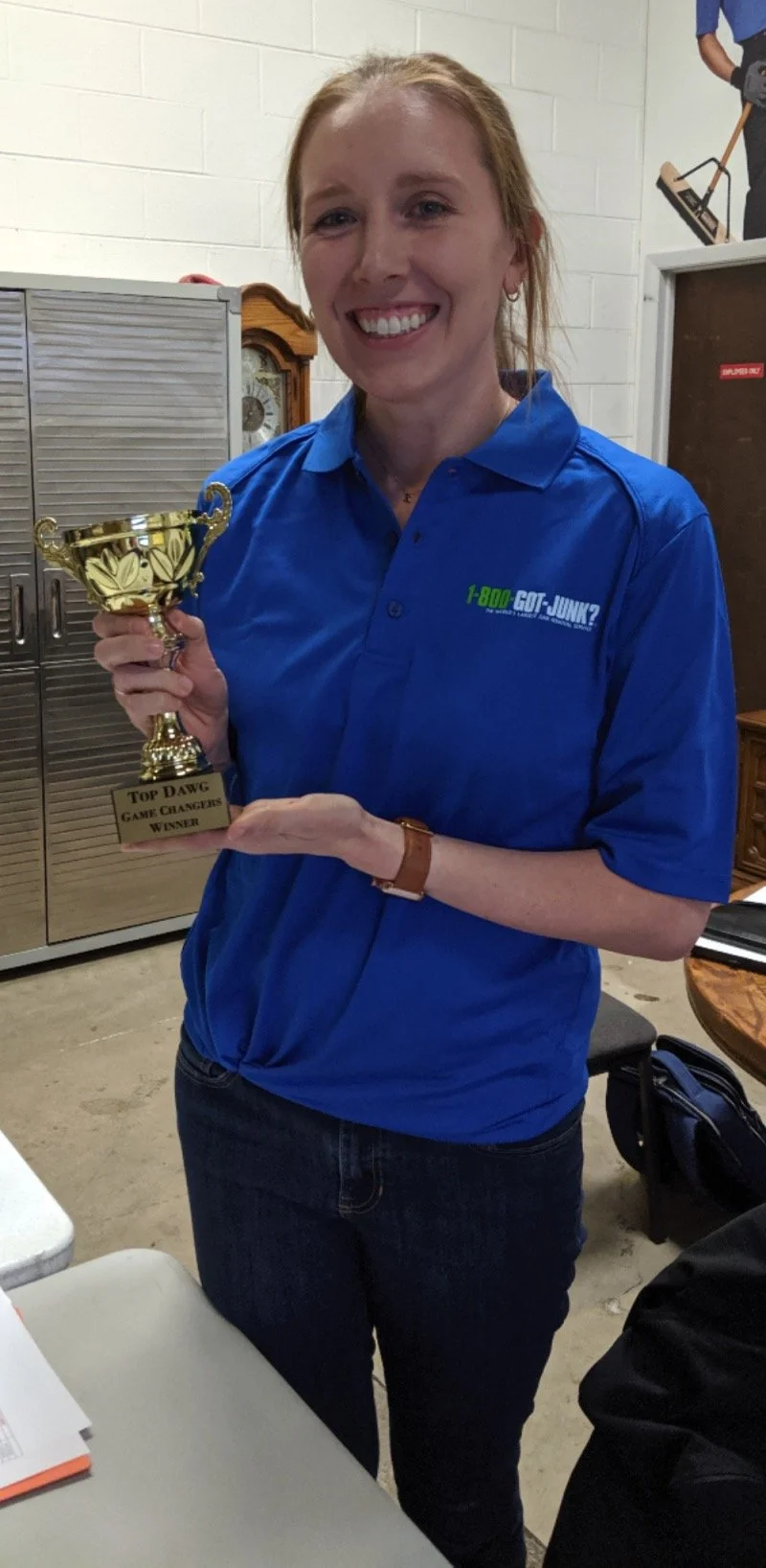 Woman smiling and holding a gold trophy cup that reads "Top Dawg Game Changers Winner," wearing a blue shirt with a logo, indoors.
