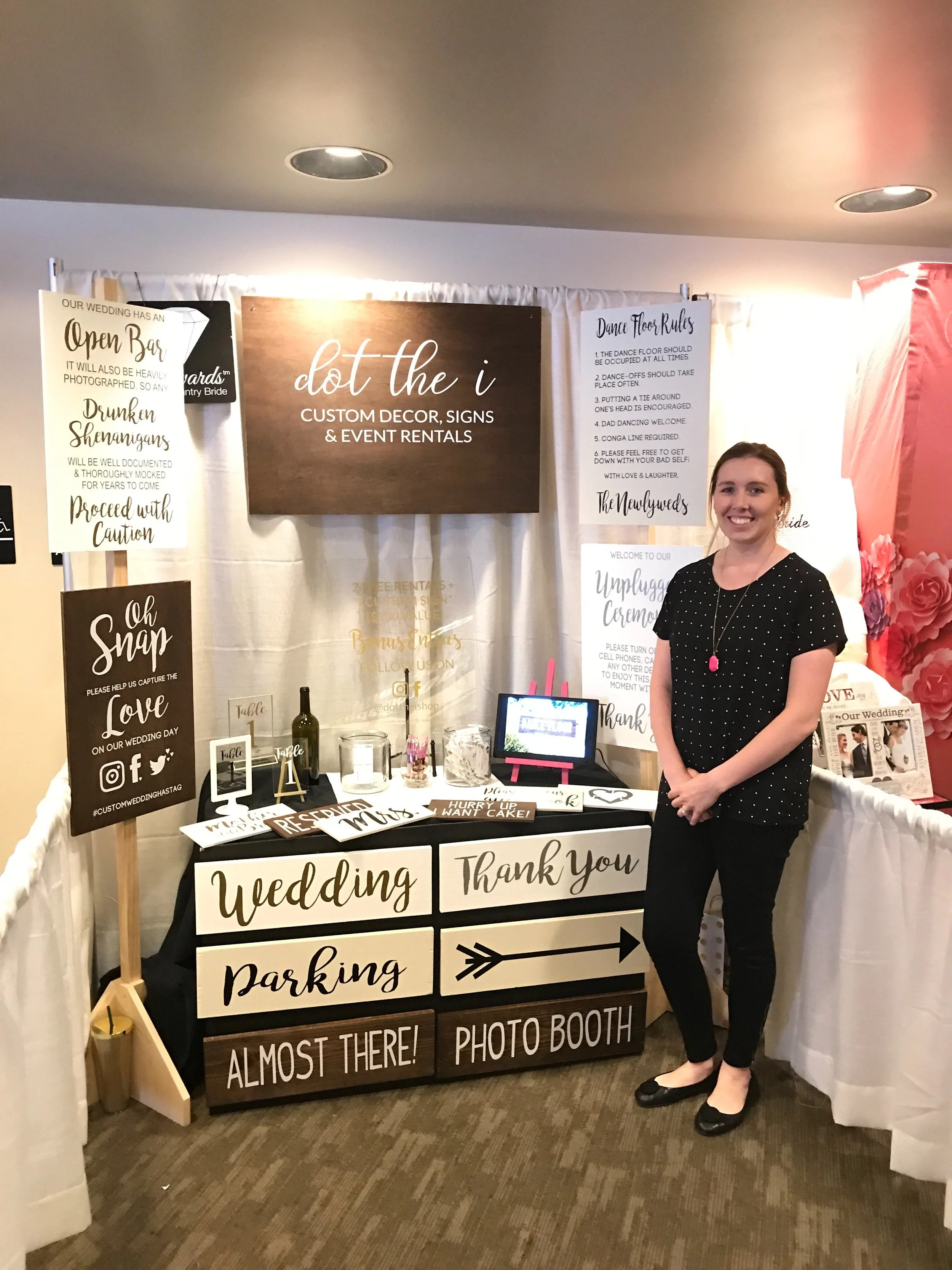 A woman stands next to a wedding sign with decor and event rental signs behind her, in a photo booth setup at a wedding or event.