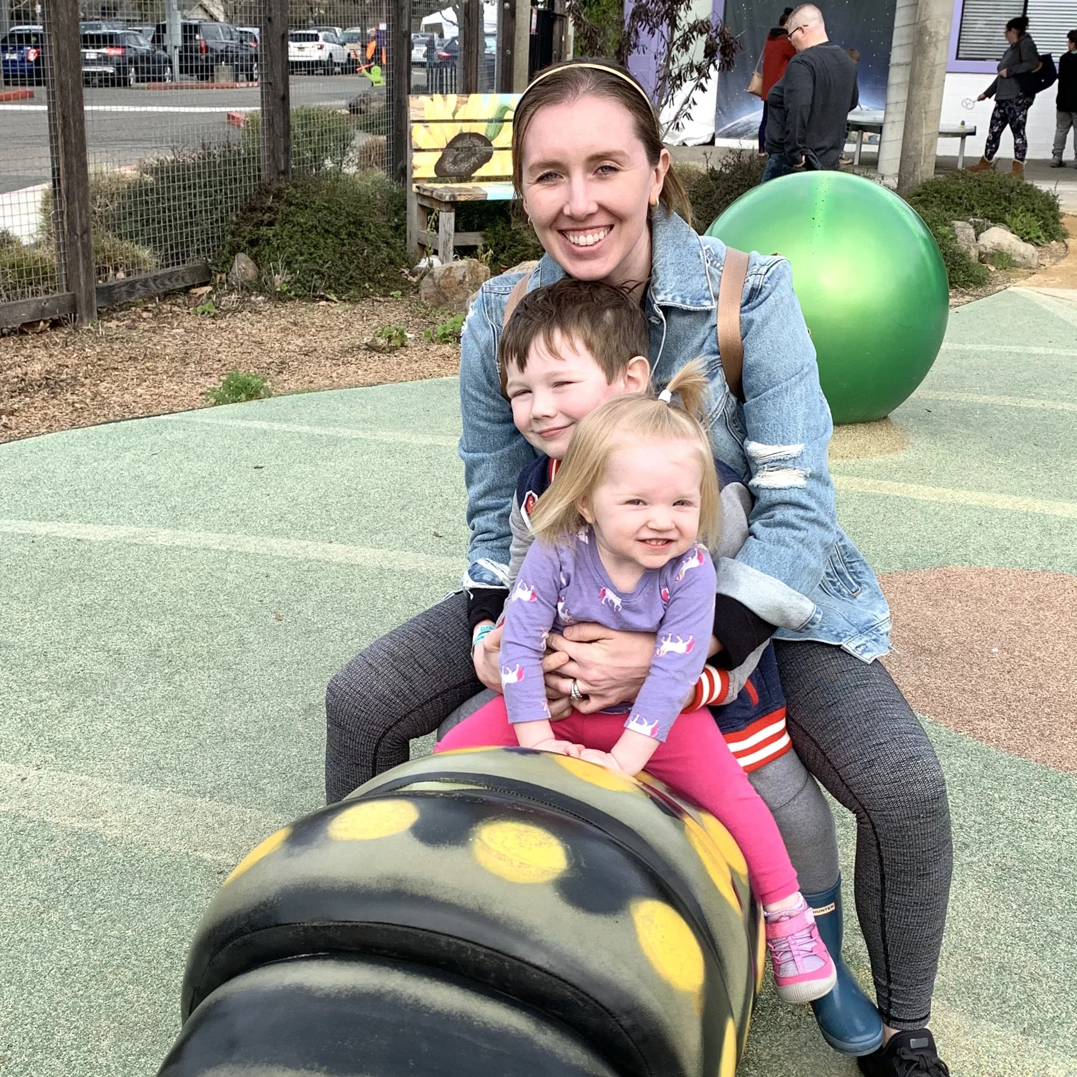 A woman smiling and crouching with three children on an outdoor playground, taken from the perspective of someone sitting on a skateboard or similar object.