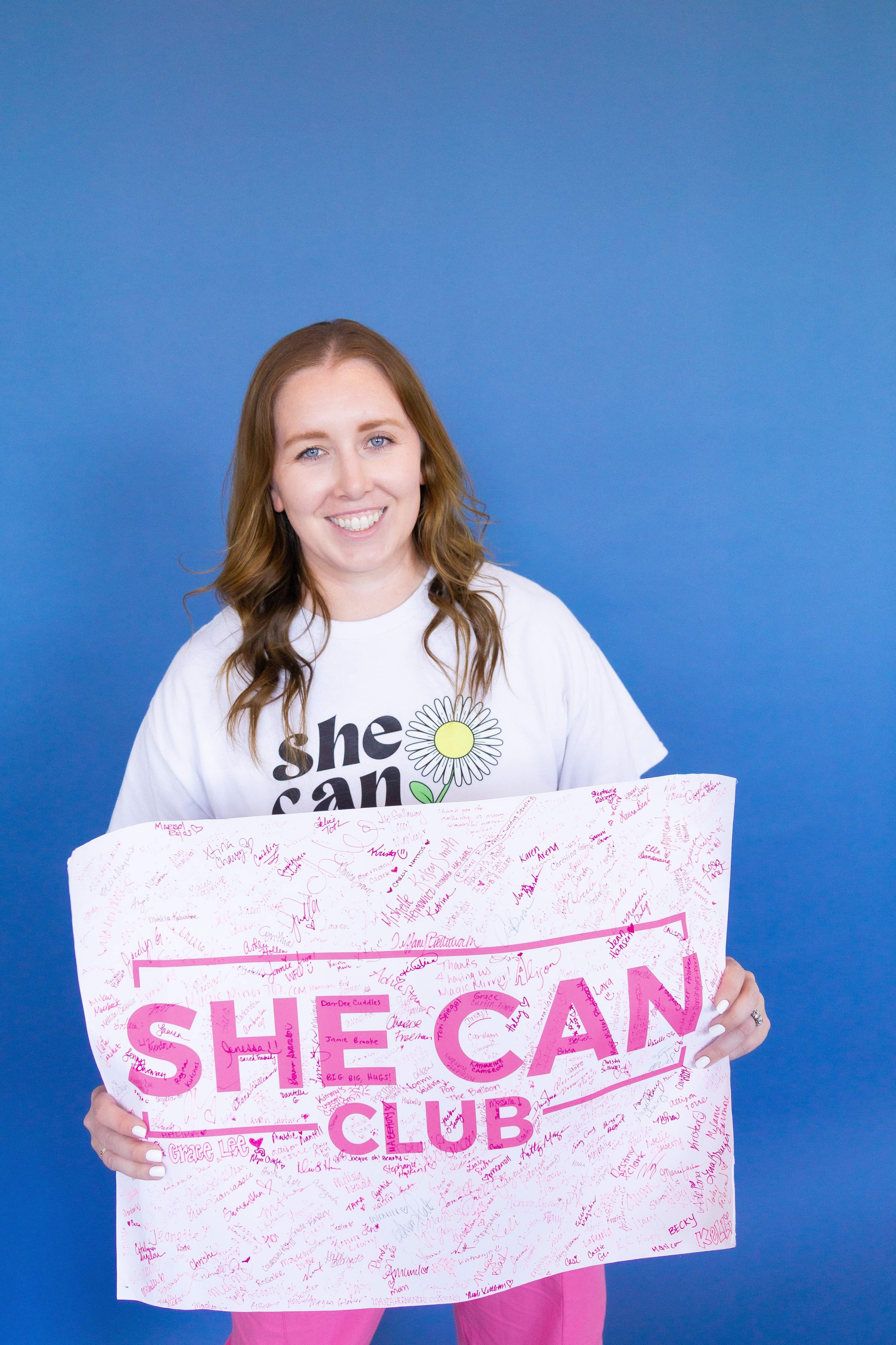 A smiling young woman with light brown hair and blue eyes holding a large white poster covered in handwritten signatures in pink ink, against a solid blue background.