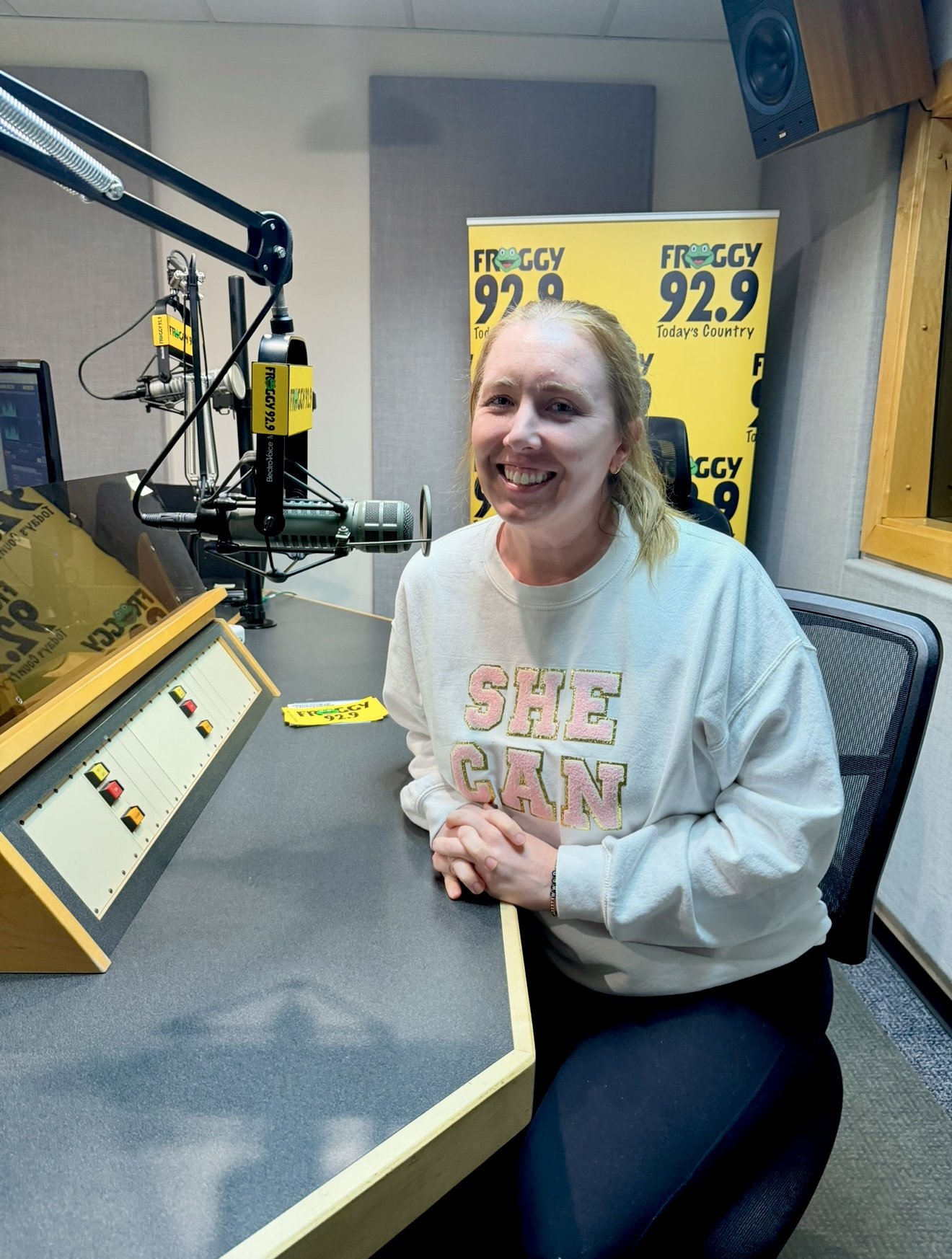 A woman sitting in a radio studio with a microphone, sound equipment, and a yellow banner behind her displaying the logo for Froggy 92.9, a country radio station.