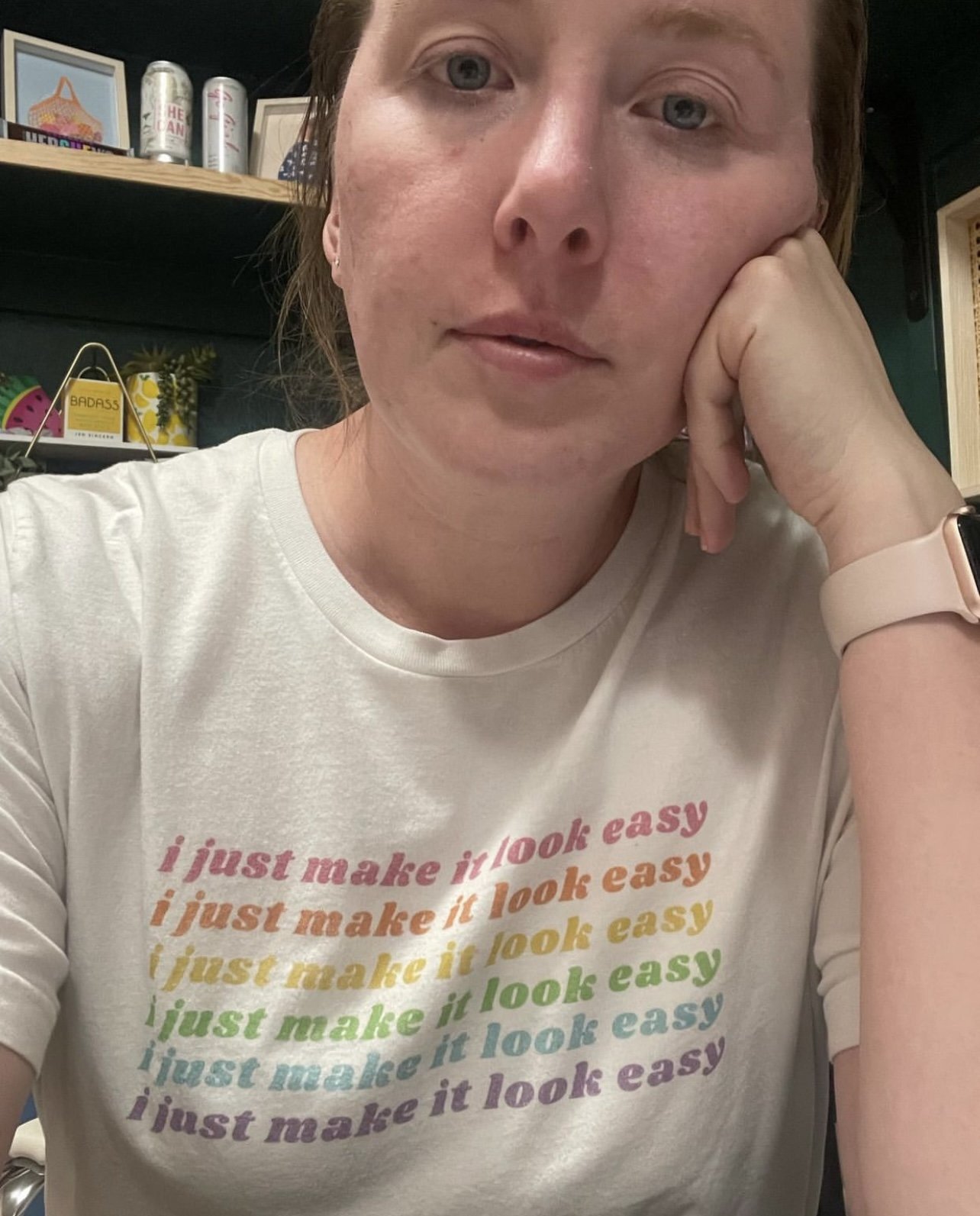 A woman with short red hair, wearing a white T-shirt with rainbow-colored text that reads 'I just make it look easy,' is resting her chin on her hand and looking at the camera.