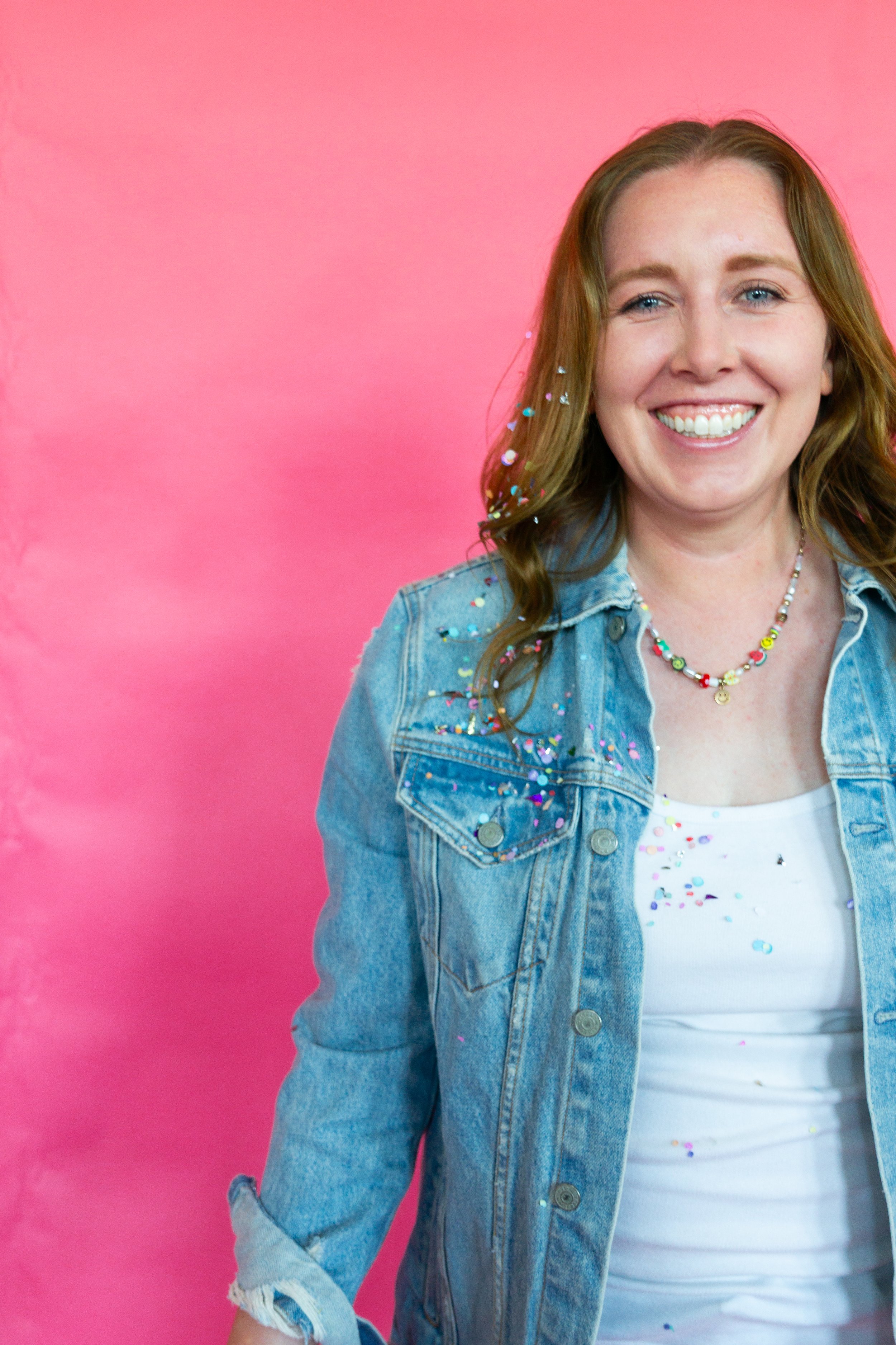 Close-up of a smiling woman with light brown hair, wearing a denim jacket, a white T-shirt with colorful confetti, and a beaded necklace, standing against a pink background.